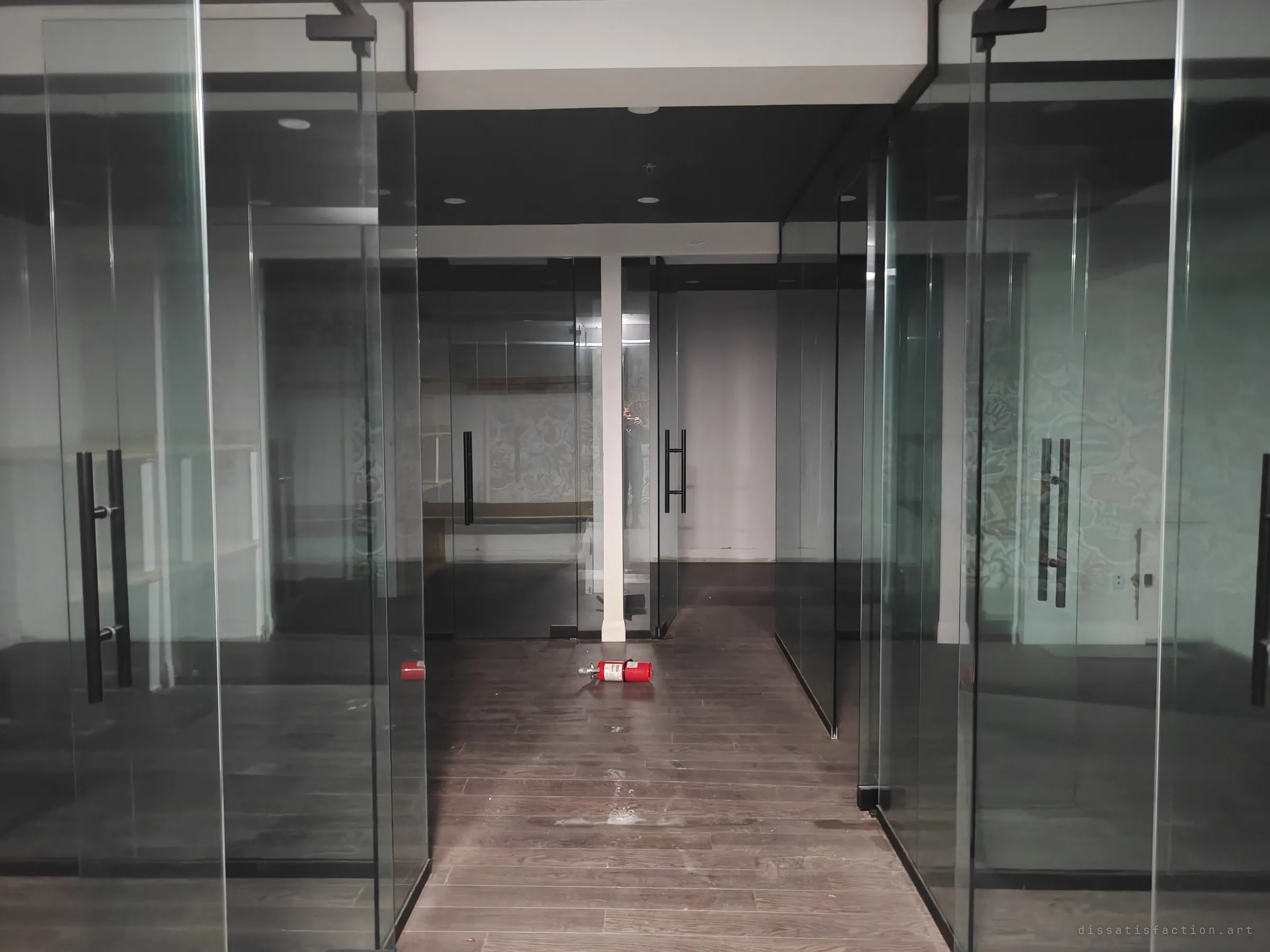 A corridor runs between glass-walled offices to a pillar at the back, a toppled fire extinguisher in the middle of the floor. Reflections stack up in the glass on both sides.