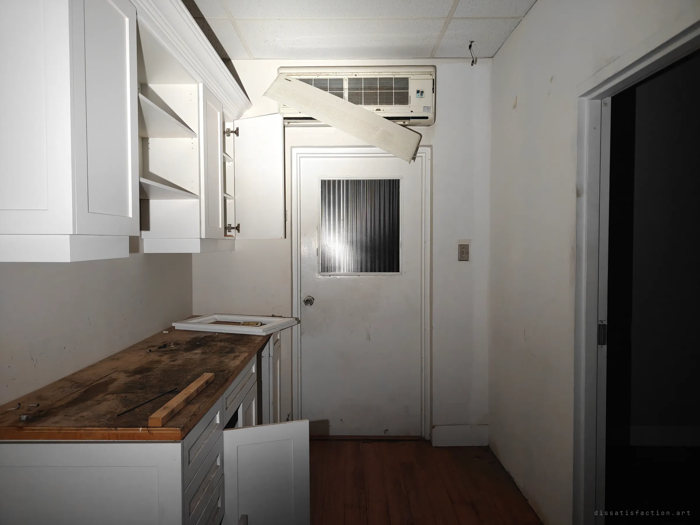 A small back kitchen with open cabinet doors, a stained worktop and a wall-mounted air conditioner whose front panel hangs loose over the door. The service rooms were left mid-strip.