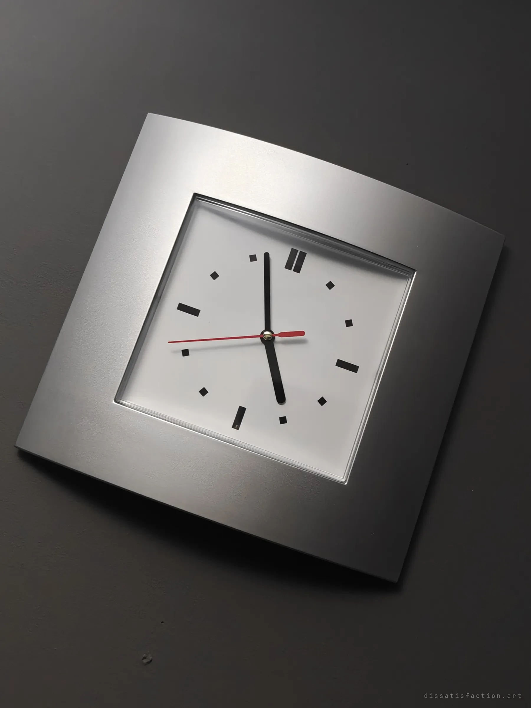 A square silver wall clock hangs askew on a dark grey wall, its hands stopped. A quiet marker of time frozen in the empty building.