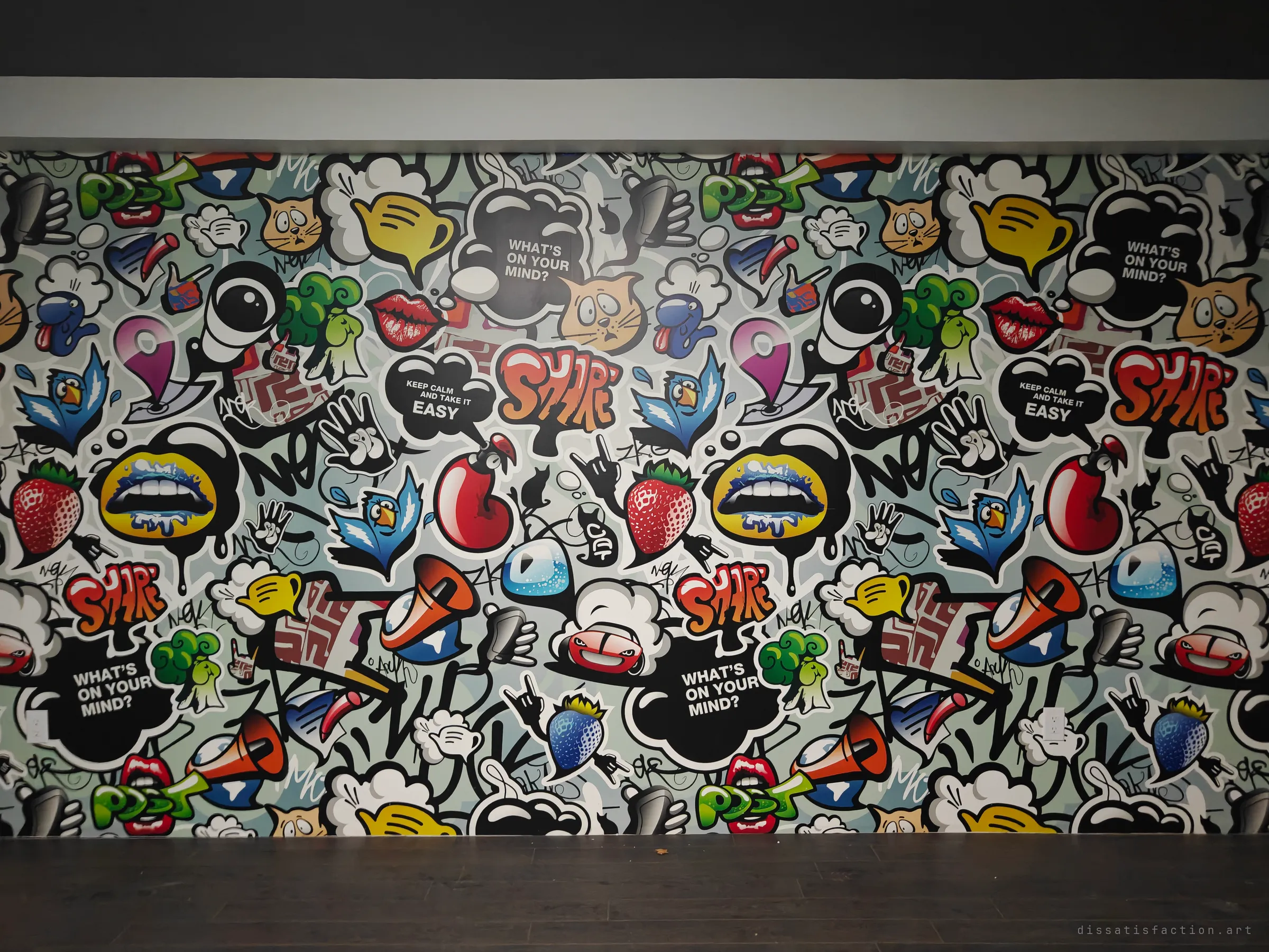 A full wall of loud cartoon-graffiti wallpaper, speech bubbles, lips and fruit, above a dark wood floor in an otherwise empty room. The bright pattern clashes with the dead silence of the space.