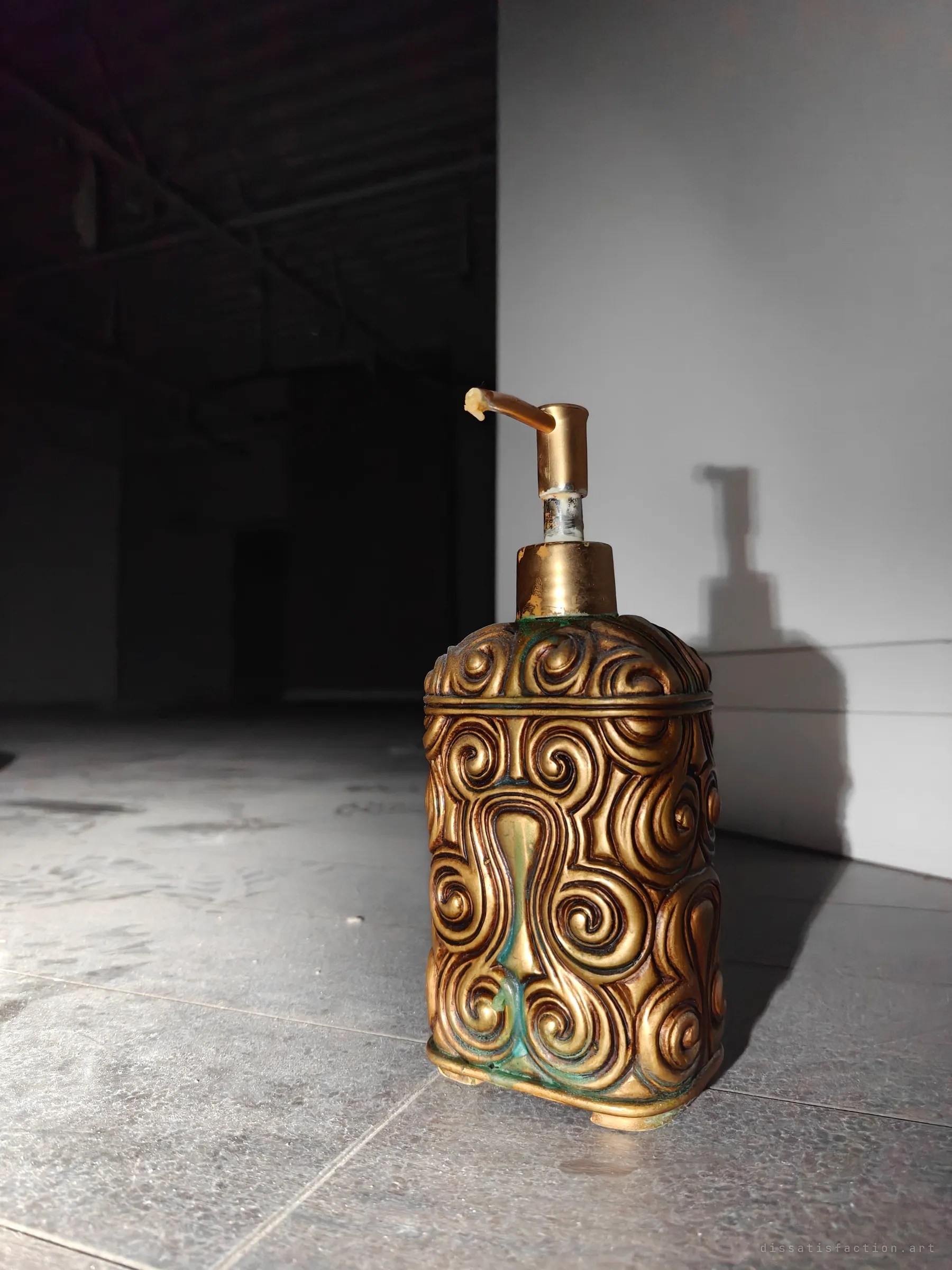 An ornate gold swirl-patterned soap dispenser stands alone on a dusty tiled floor, lit hard from the side against a dark storeroom. A small decorative object left behind in a bare space.