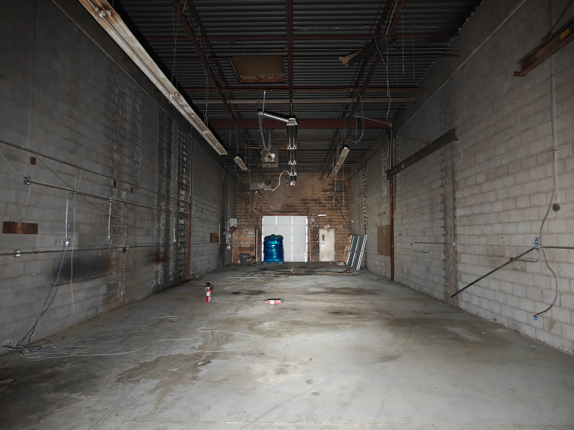 A long, empty cinder-block warehouse bay with exposed steel roof decking, dangling wires and fire extinguishers on the stained concrete floor, leading to a roll-up loading door. Shows the industrial back-of-house scale of the building.