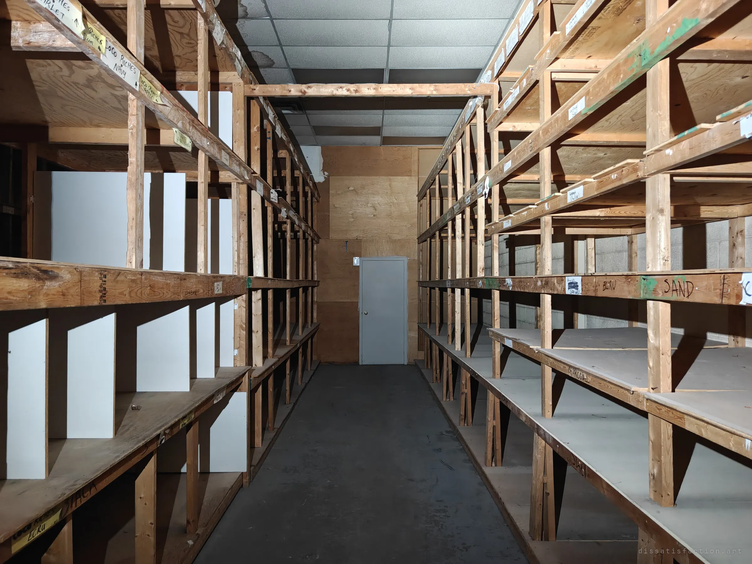 An aisle of tall handmade wooden shelving, empty except for taped labels naming fabric colours, leading to a grey door. The labels hint at the stock that once filled every bay.