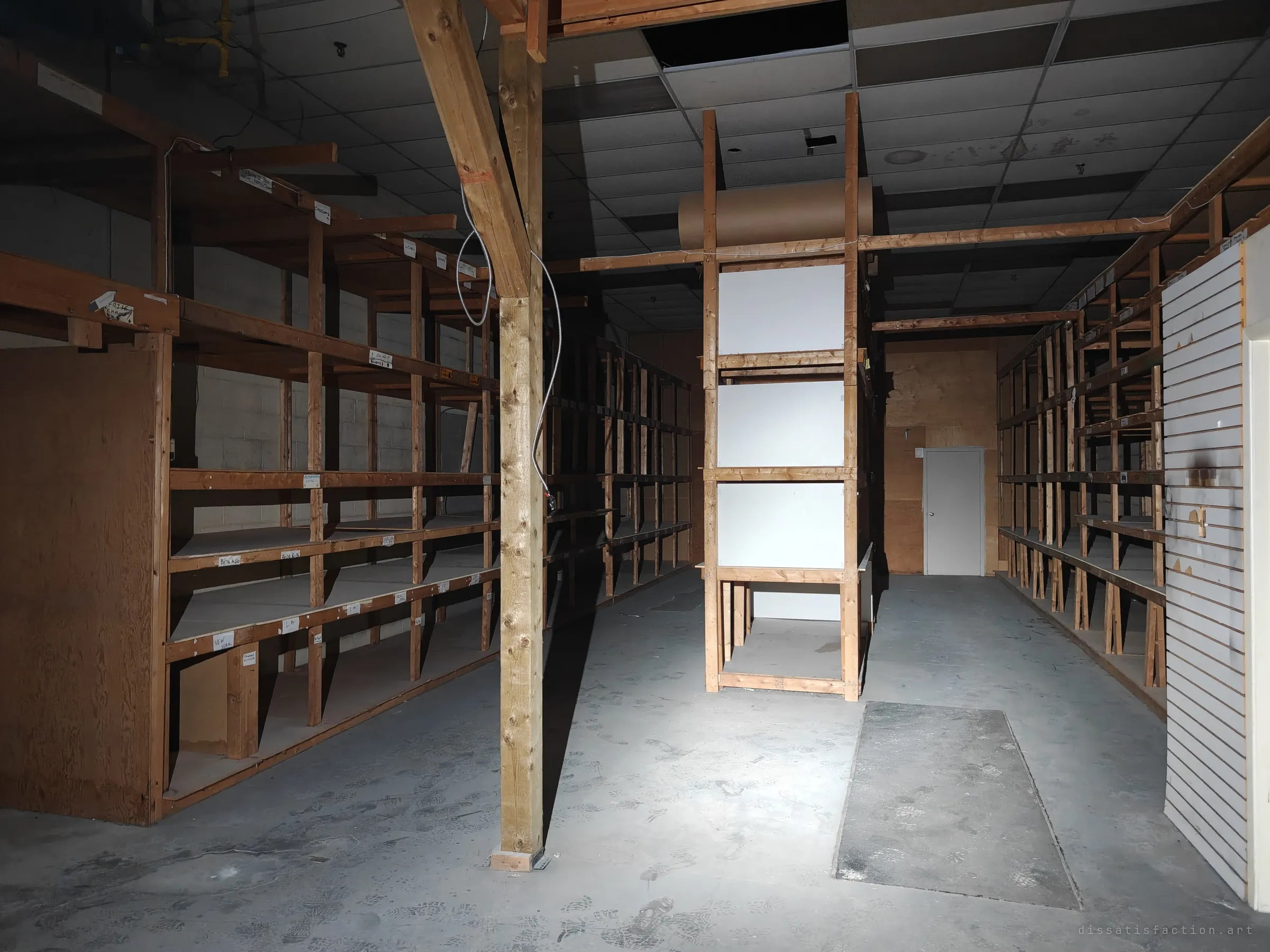 A dark stockroom of empty timber shelving bays around a central post, with a roll of brown paper on top and a scuffed concrete floor. The rows of bare racks show the full size of the old inventory.