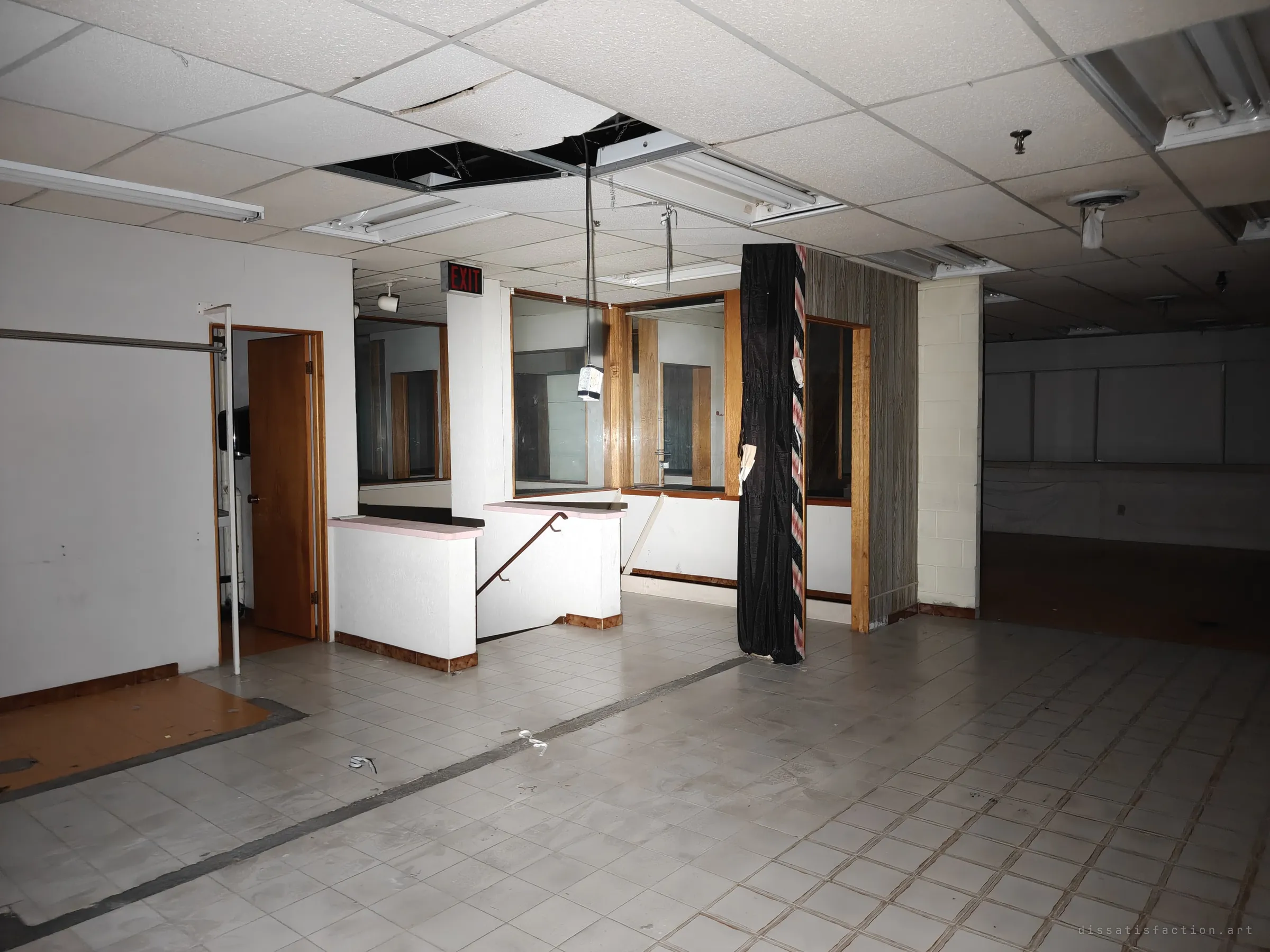 An open retail floor with sagging ceiling tiles, dangling light fixtures, an EXIT sign and a stairwell railing behind low partition walls. The bare tiled floor still shows the outline where counters once stood.