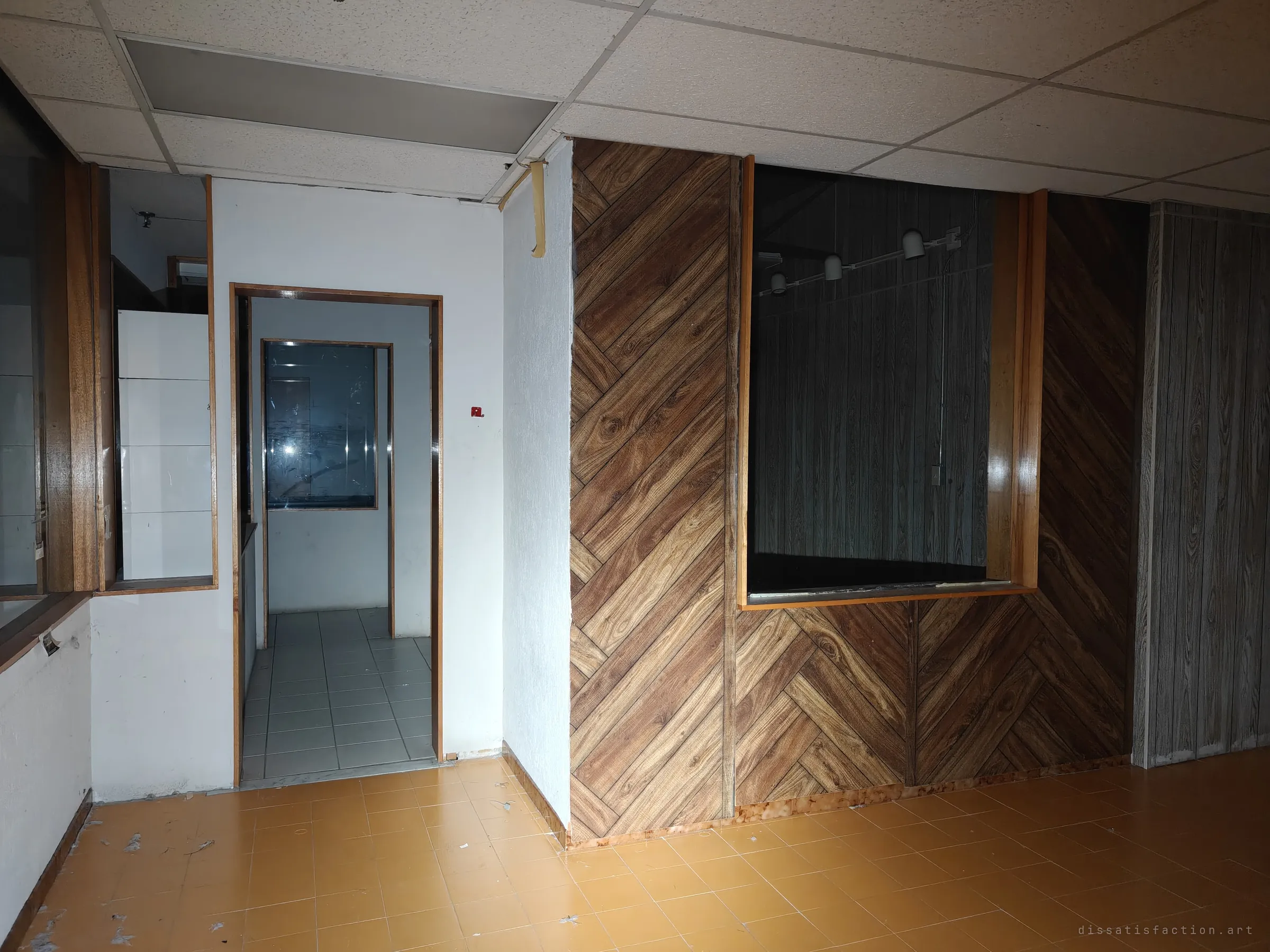 An emptied office corner with herringbone wood-look panelling, a dark service window and a doorway into a white tiled back room. The dated finishes set the showroom's late-century mood.