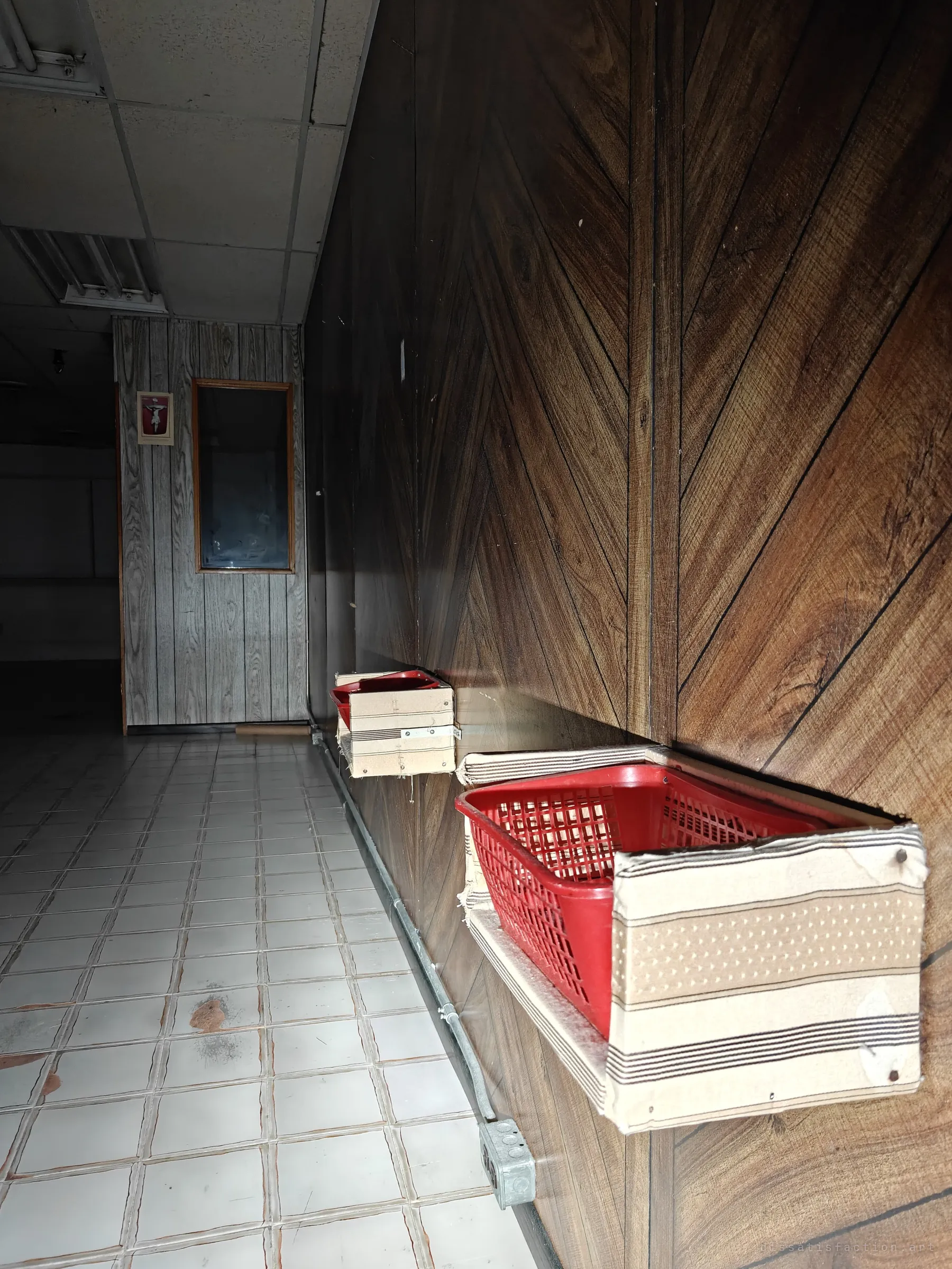 Red plastic baskets still hang in fabric-wrapped holders along a wood-panelled hallway, with a small crucifix on the door at the far end. Left-behind fittings hint at the staff who once worked here.