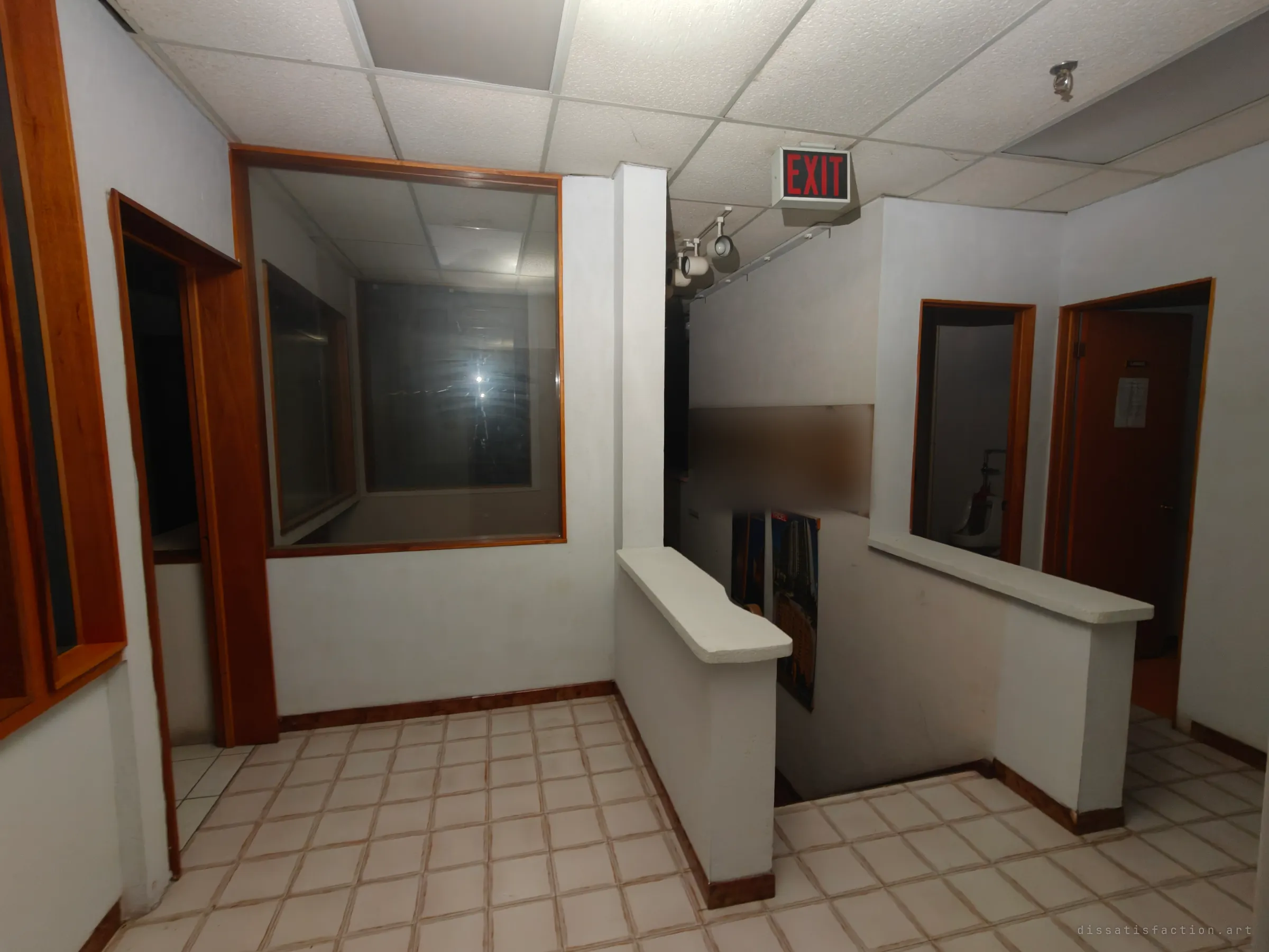 An upstairs landing with half-walls around a stairwell, an exit sign and a faded promotional banner and posters on the stair wall. Glass-fronted offices open off the landing.