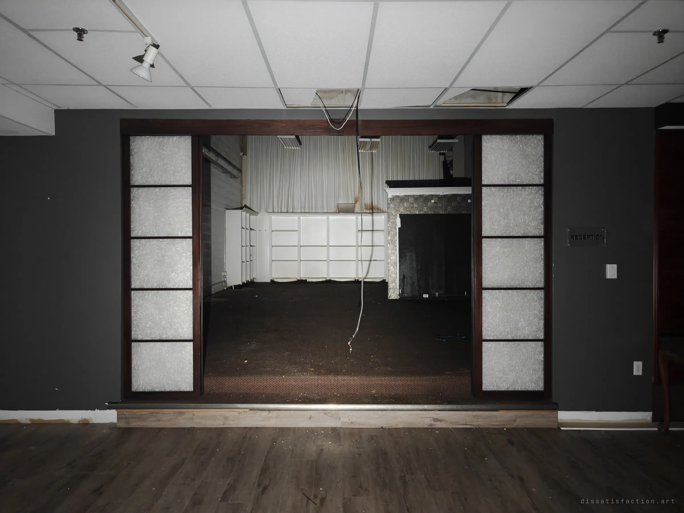 A reception room with dark walls and frosted shoji-style panels frames a wide opening into a gutted showroom, with a loose cable hanging from the torn ceiling. The symmetry makes the emptiness feel staged.