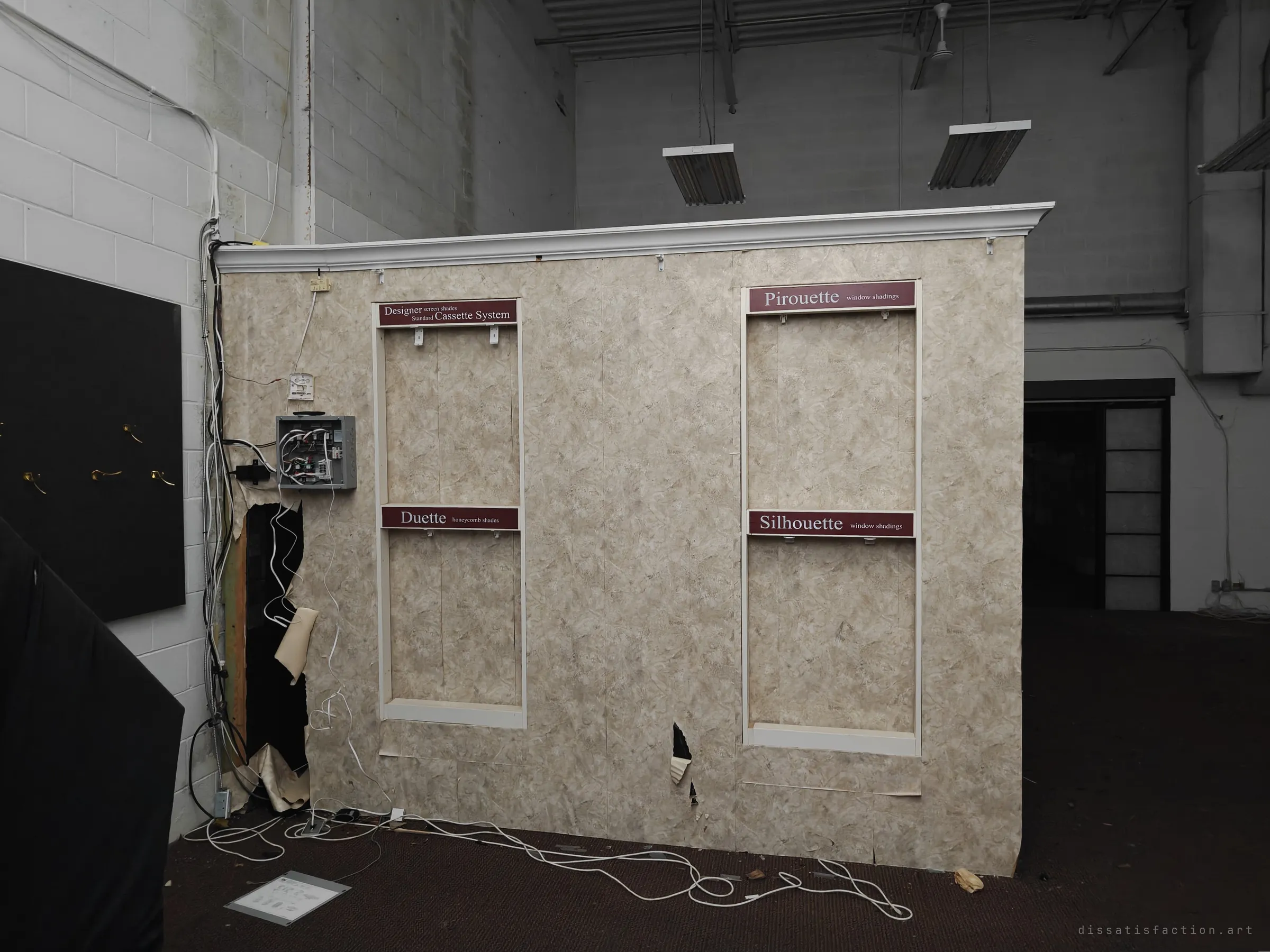 A freestanding display wall in a high warehouse space still carries labels for window-shade product lines, but the blinds themselves are gone and cables spill across the floor. It shows the business stripped down to its signage.