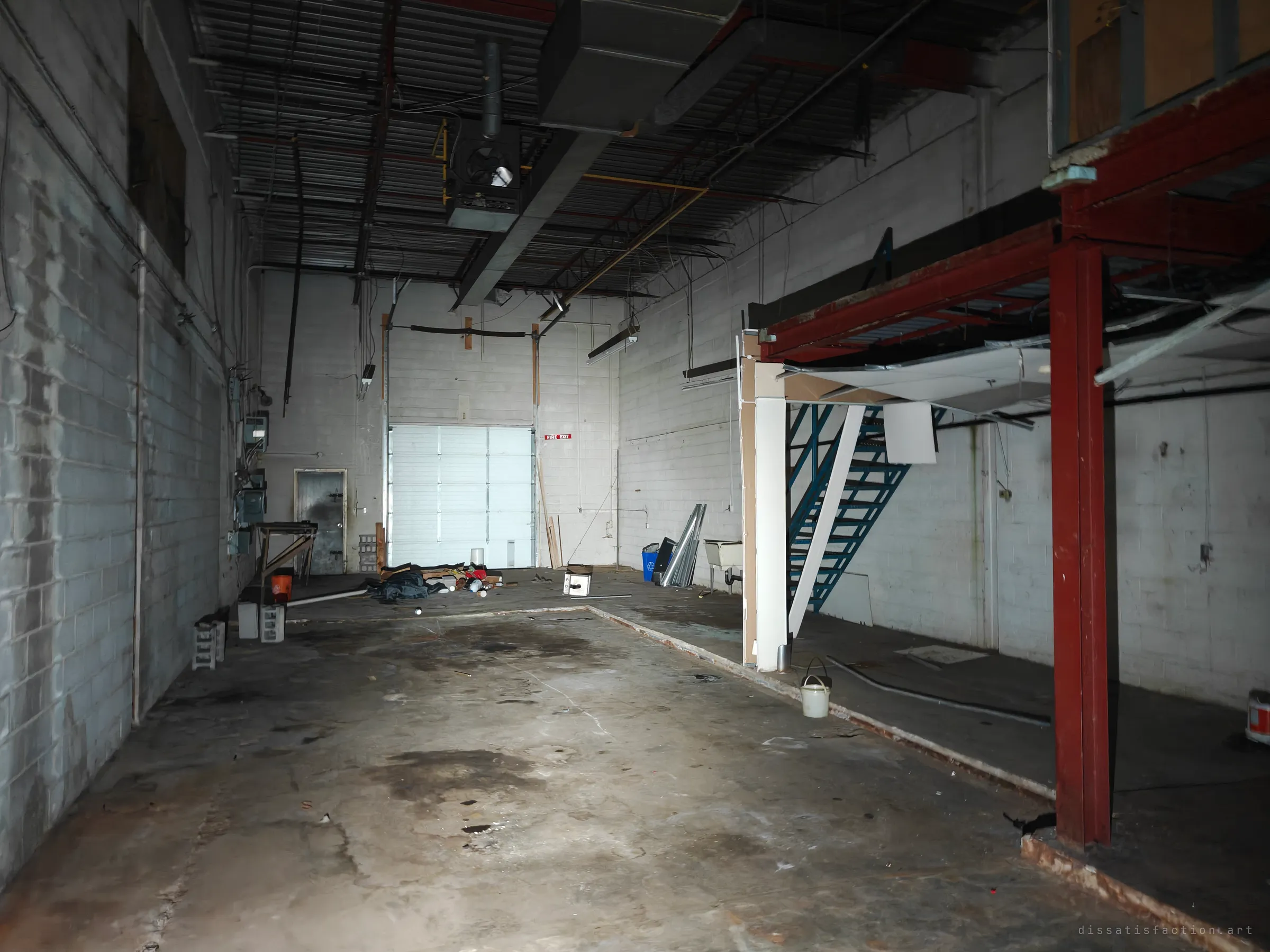 A long cinder-block warehouse bay with a roll-up loading door at the far end, a red steel mezzanine and a blue staircase on the right. Debris and stains cover the concrete floor.