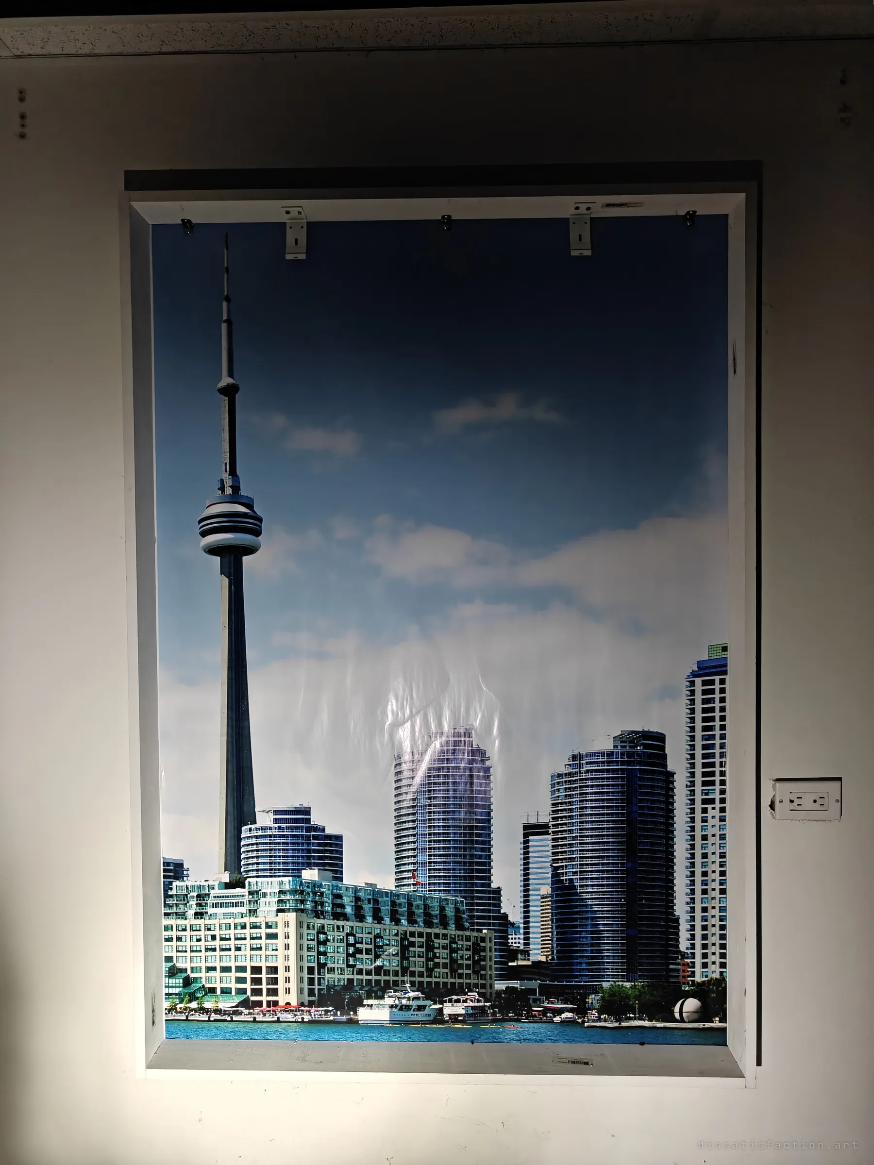 A fake window lit by a flashlight holds a printed photo of a city skyline and waterfront, its plastic sheet sagging. A showroom sample for window treatments, now left in the dark.