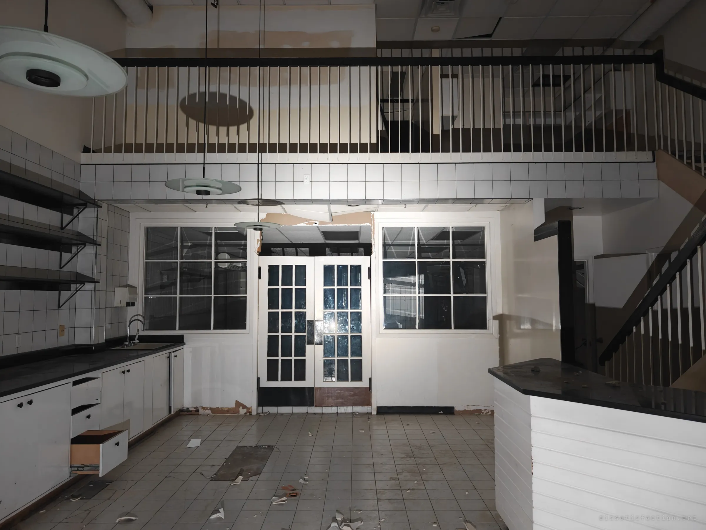 A double-height former cafe space with white tiled walls, black countertops and an open drawer, under a railed mezzanine. Glass-paned doors lead to a back room, the place stripped but its fittings still in place.