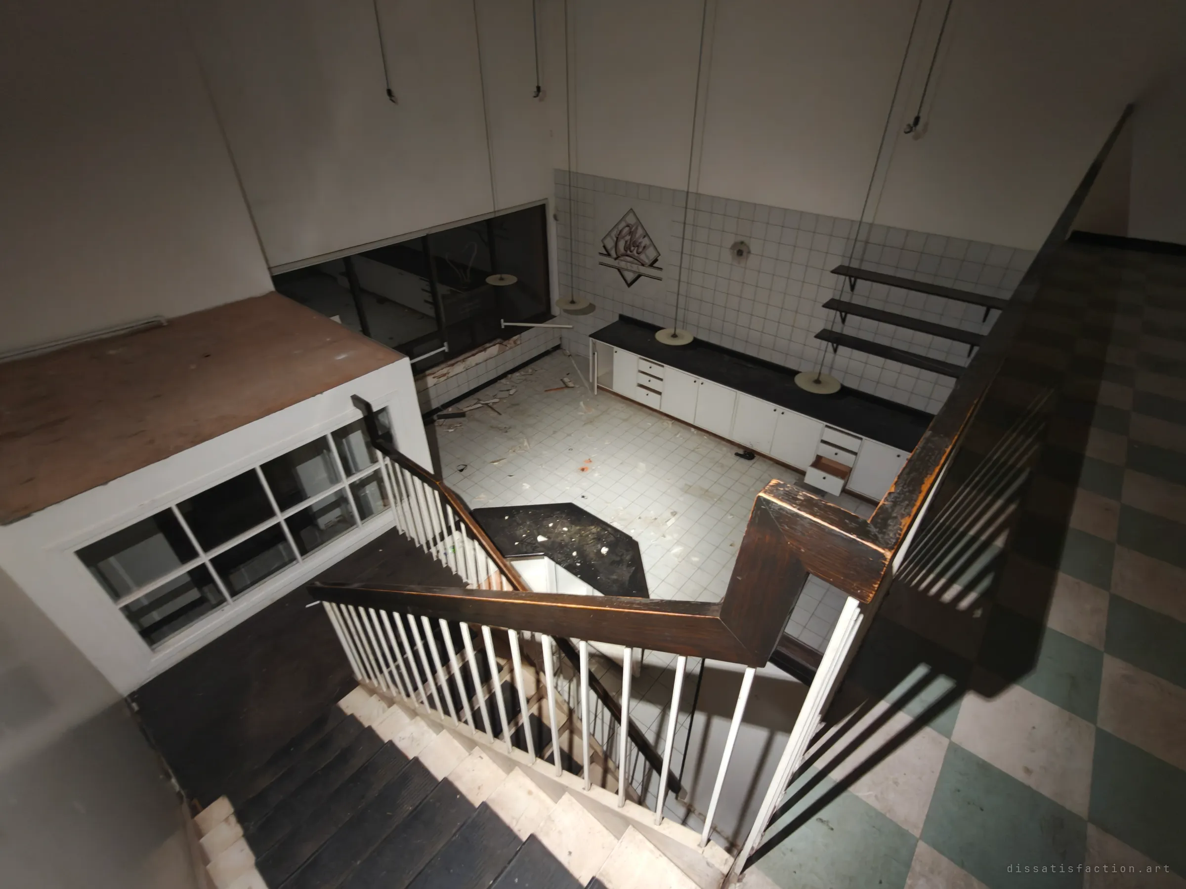 Looking down the stairwell from the mezzanine into the same tiled cafe below, with its hanging disc lamps and debris-strewn floor. The high angle shows how the upper floor wraps around the old service area.
