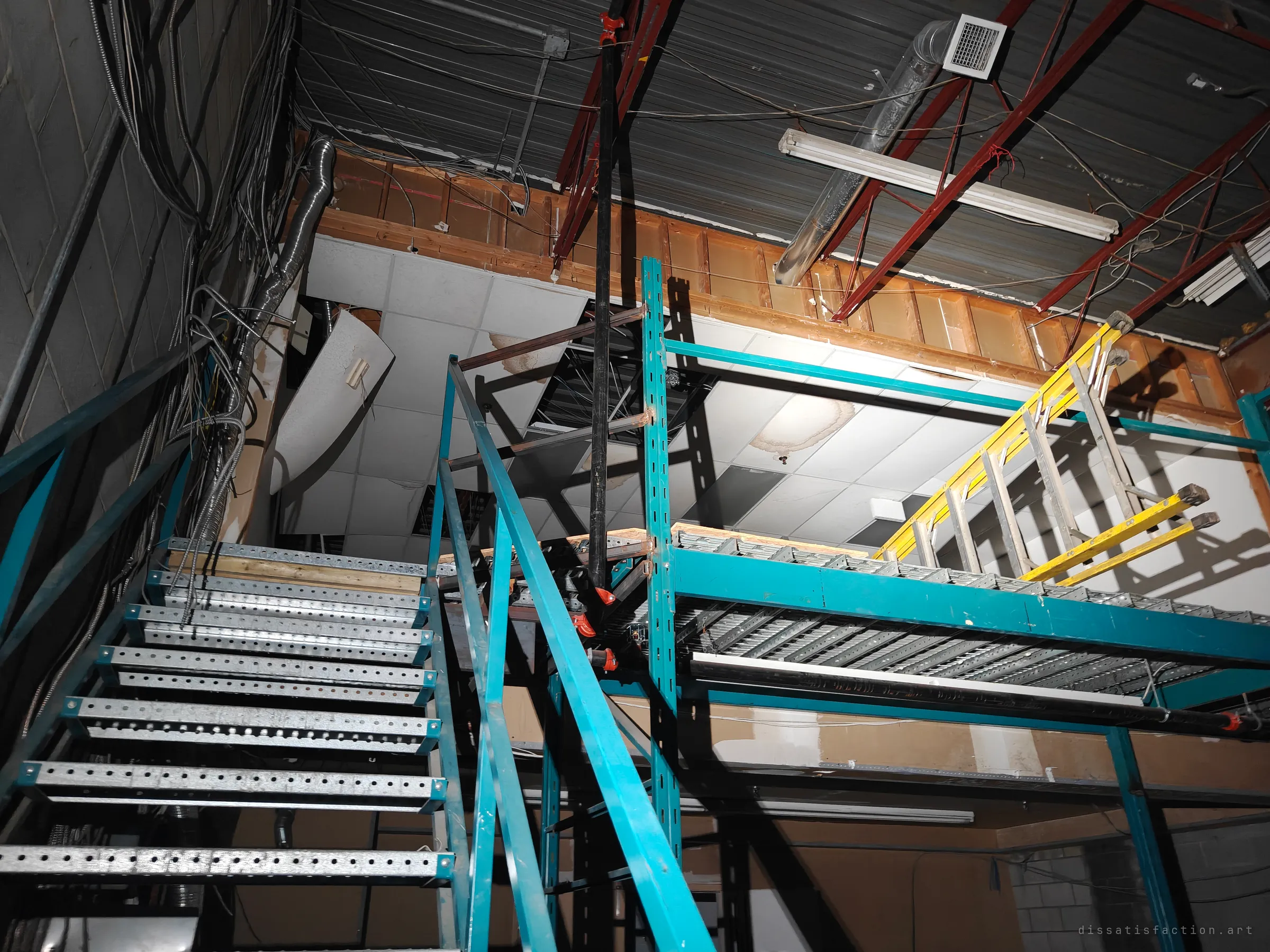 A makeshift steel stair and teal pallet-rack mezzanine climb into an exposed ceiling of red joists, loose cables and collapsed ceiling tiles. A yellow ladder is left lying on the upper deck.