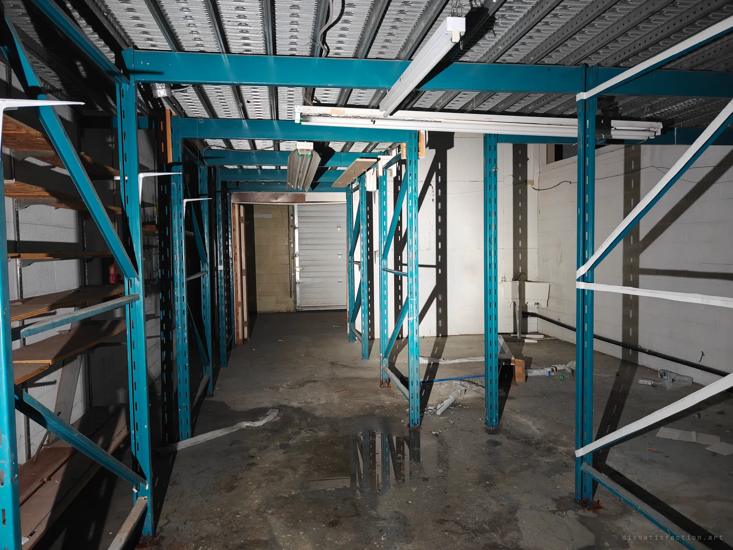 Beneath the teal racking mezzanine, empty bays run back toward a closed roll-up door, with a puddle on the concrete floor reflecting the uprights. It reads as a stockroom emptied out and left to the damp.