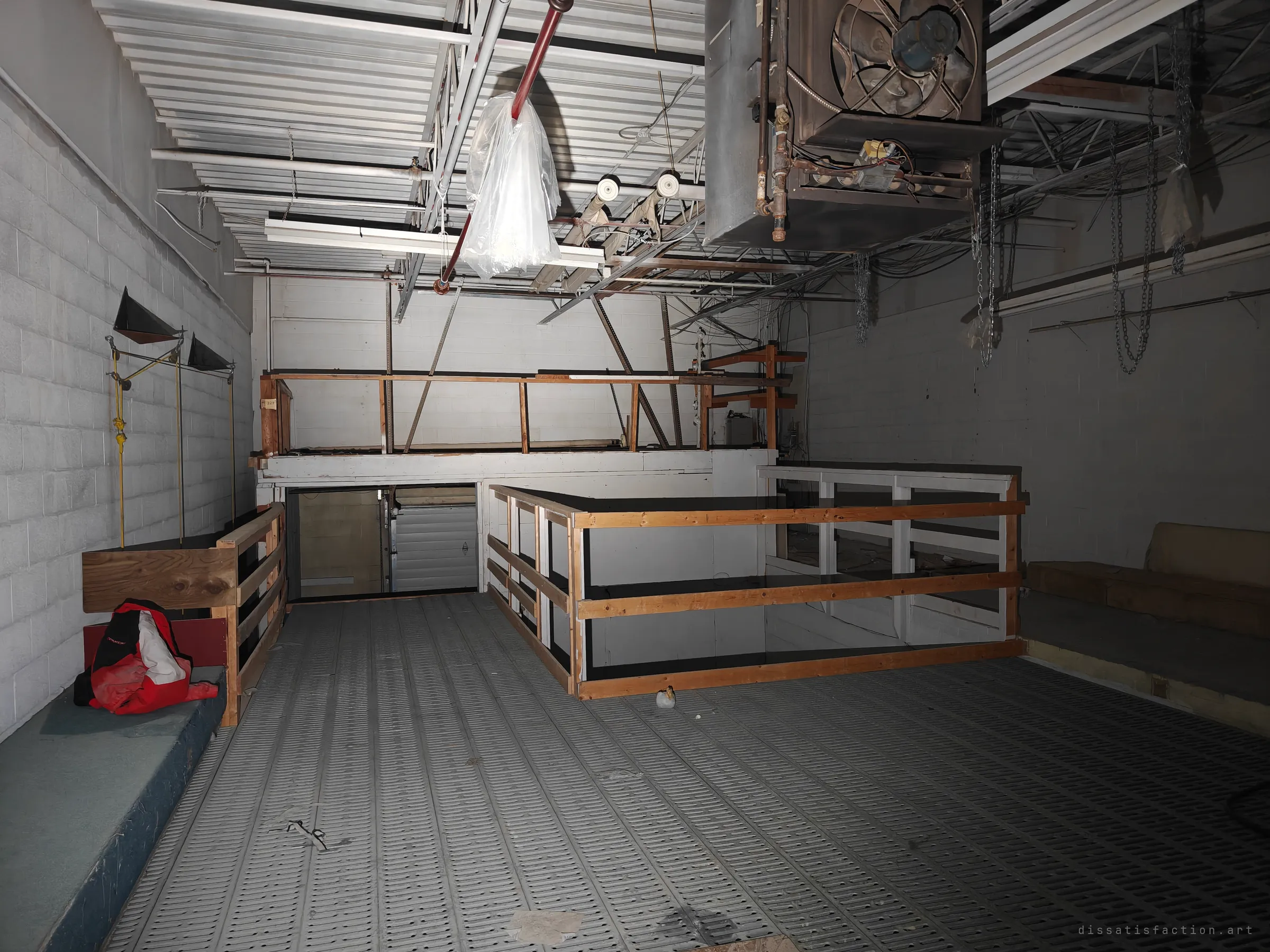 An upper storage deck with a grated floor and timber railings around a stair opening, a large unit heater hanging from the roof trusses. A red bag and scattered junk are the only things left behind.