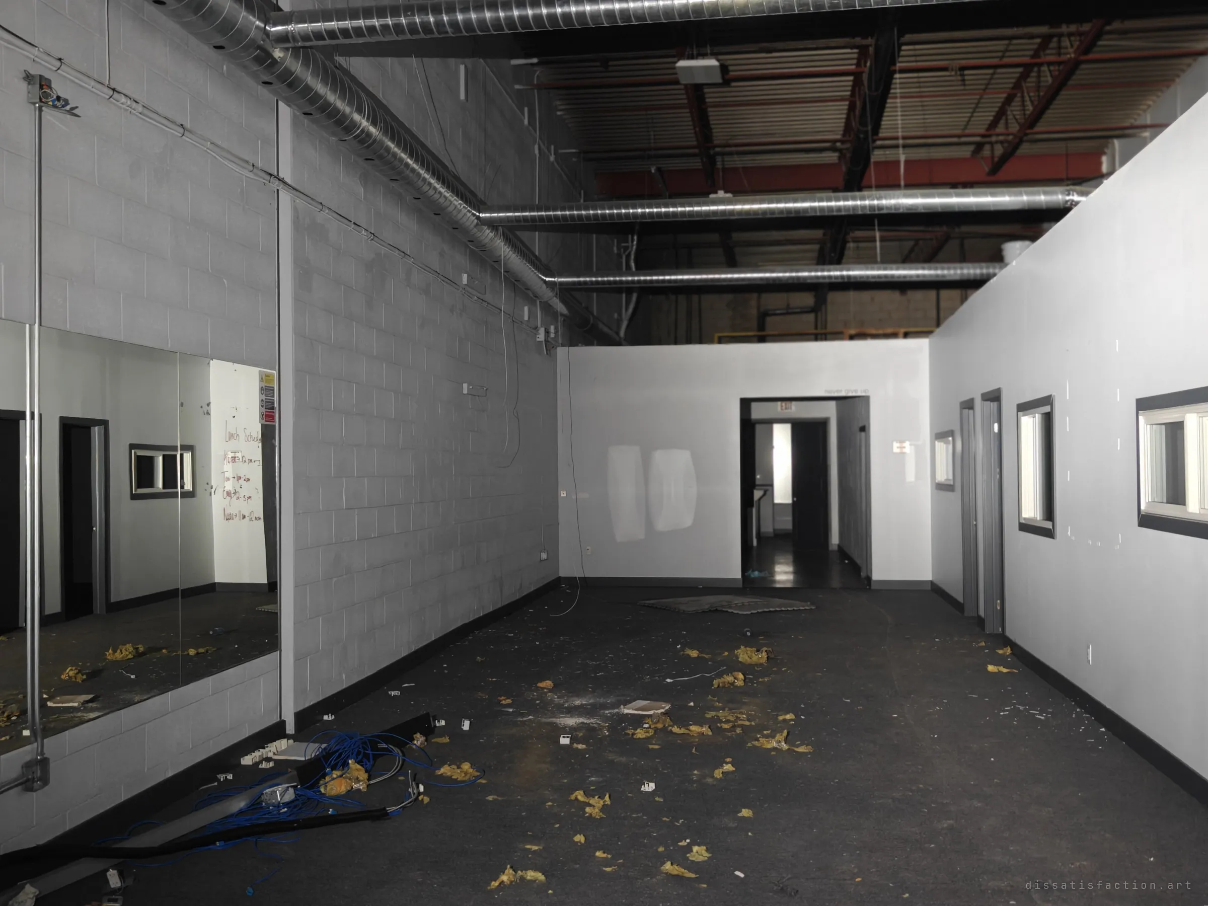 A long grey studio room with a mirrored wall, black floor and silver ducting overhead, strewn with insulation and loose network cable. Small office windows line the right-hand wall toward a lit doorway.
