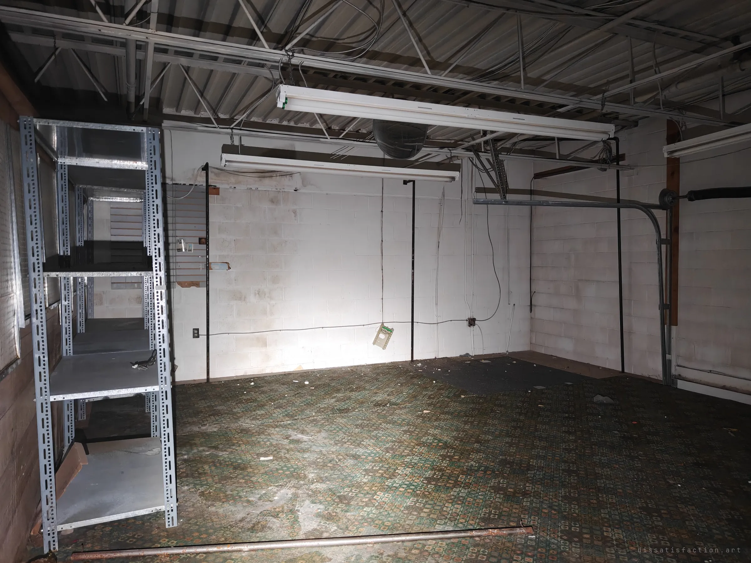 A bare cinder-block storeroom with a patterned green carpet, a galvanised shelving unit and a roll-up door track on the right. Fluorescent fixtures still hang from the open steel roof.