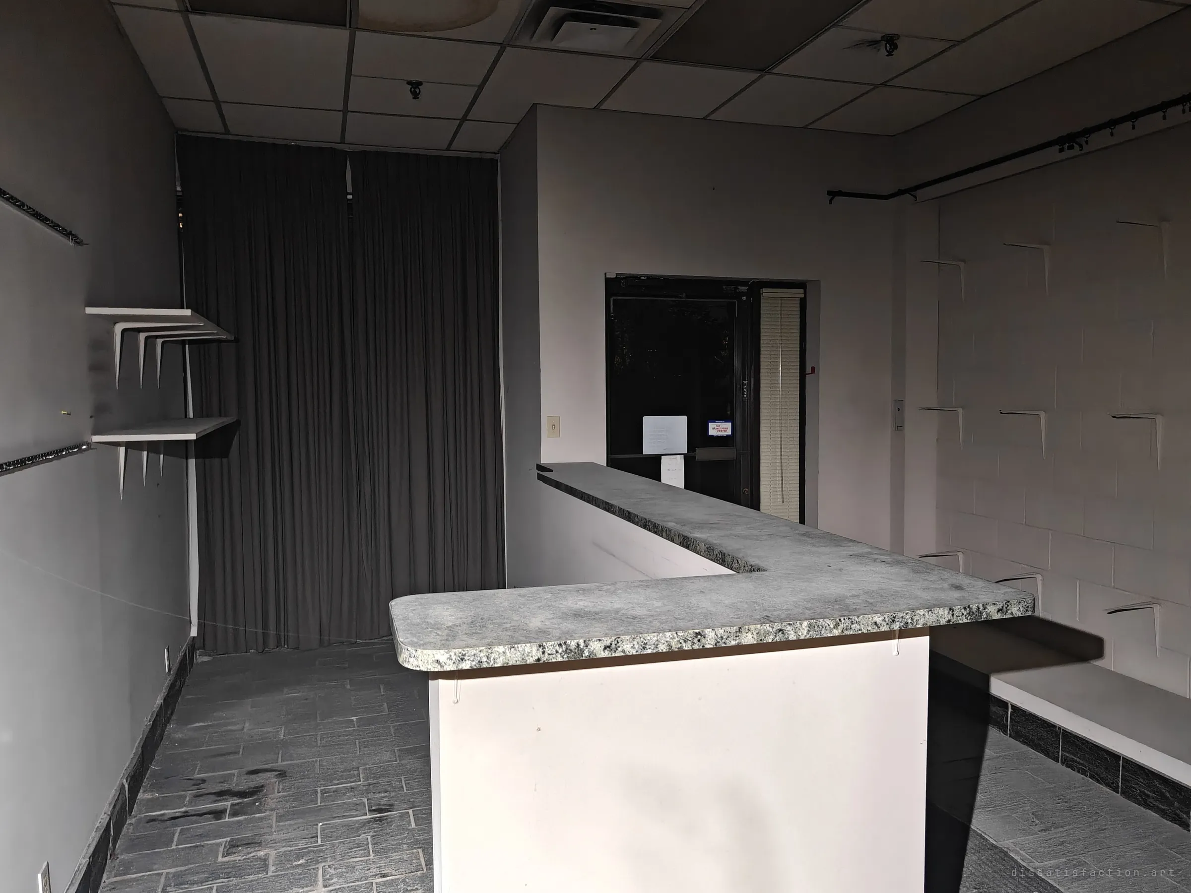 A small shopfront unit with a speckled granite counter, empty shelf brackets on the walls and a heavy curtain at the back. Notices are still taped to the dark glass entry door.