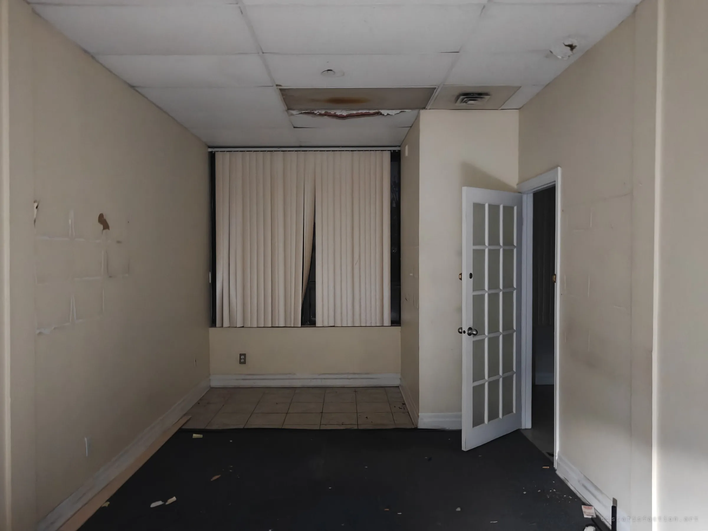 A narrow beige office with a stained drop ceiling, vertical blinds drawn over the window and a French door standing open to a dark hallway. Paint scars on the wall mark where things once hung.