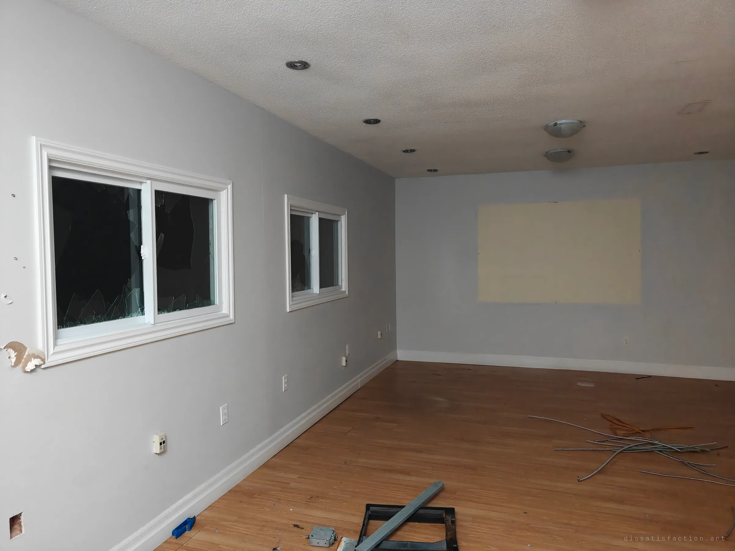 A long, emptied room with grey walls, smashed sliding windows and a pale rectangle where a screen once hung. Bundles of stripped conduit lie across the wood floor, the leftovers of copper scrappers.