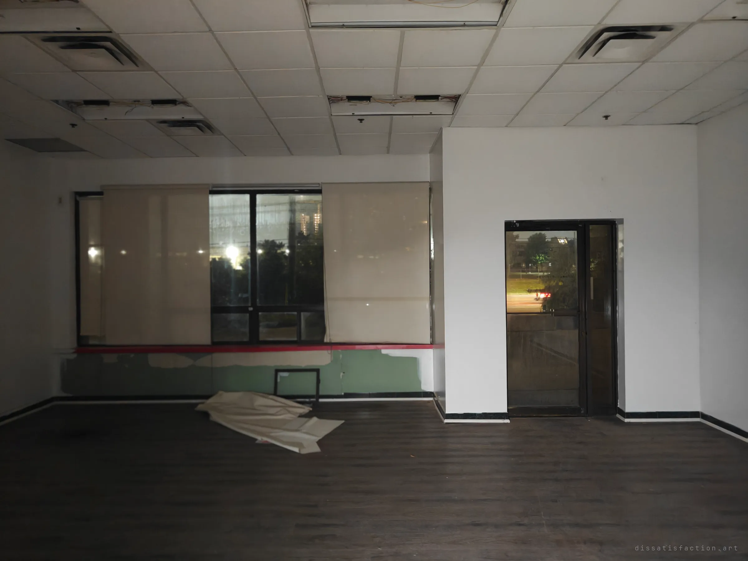 A dark former storefront with a dropped tile ceiling, gutted light fixtures and a torn blind lying on the floor. Through the window and the glass door, streetlights and passing cars show the city carrying on outside.