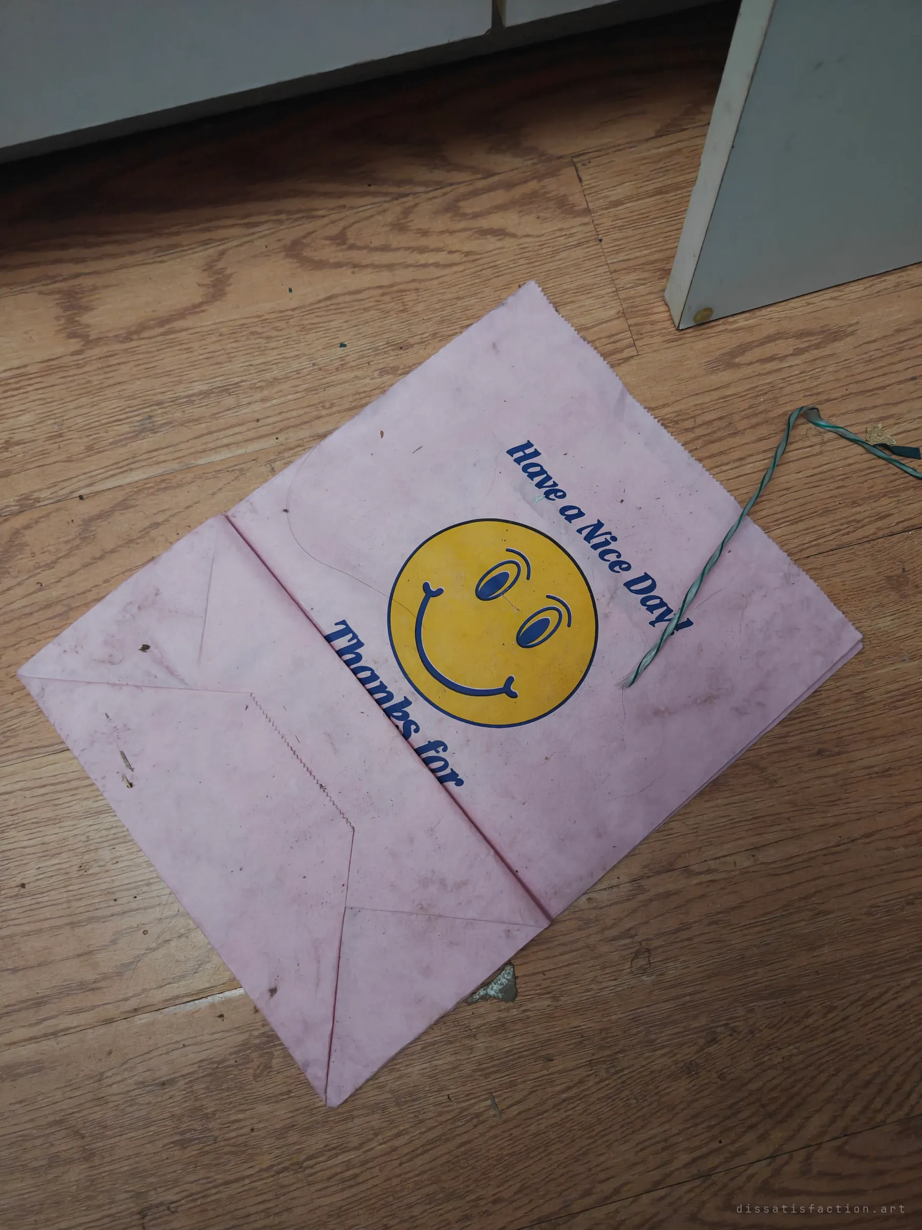 A flattened pink takeout bag with a yellow smiley face wishing a nice day, dusty on a wood floor. A cheerful leftover in an abandoned space.