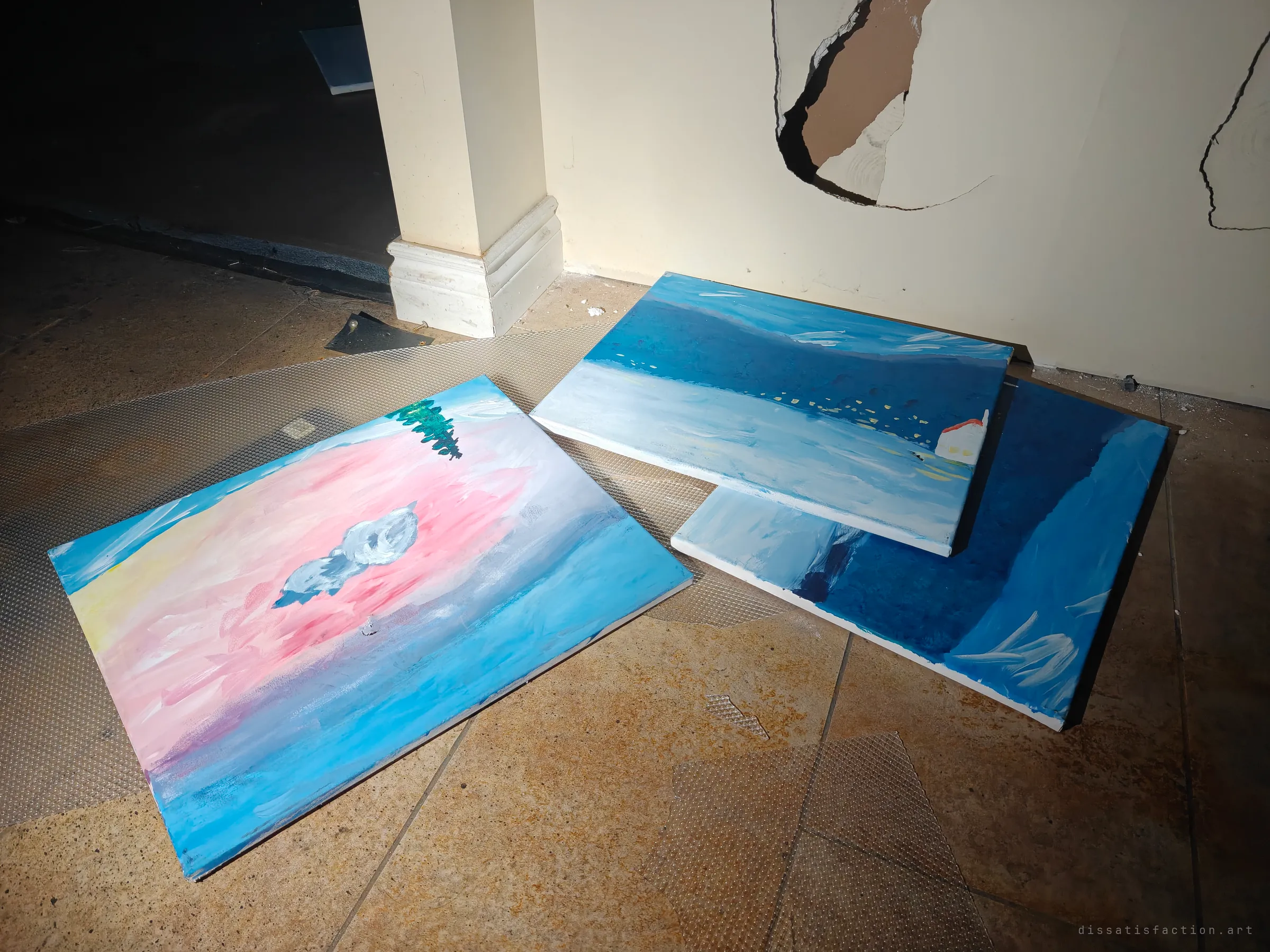 Three amateur paintings of pink and blue skies and water lie abandoned on a tiled floor below a wall with holes punched through the drywall. Someone's hobby art, left behind in the dark.