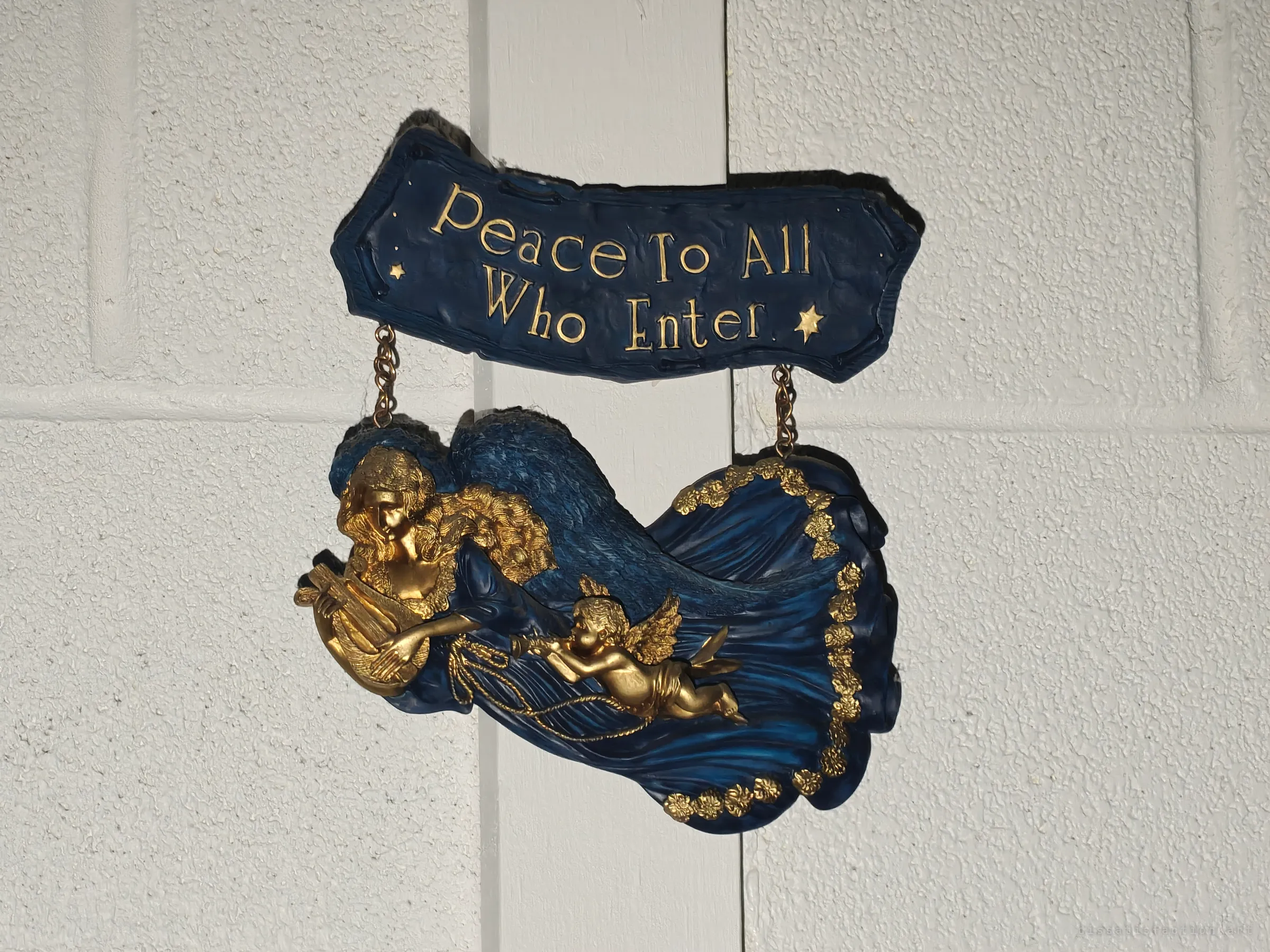 A blue and gold resin angel with a cherub hangs on a white block wall under a banner reading "Peace To All Who Enter". The welcome still hangs in a building no one is meant to enter.