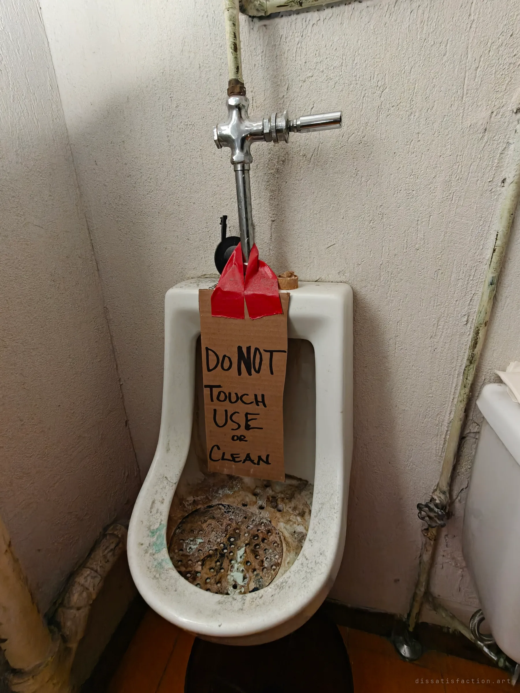 A grimy urinal with a corroded drain and a cardboard sign taped to its flush pipe warning not to touch, use or clean it. A blunt handwritten order from the building's last days.