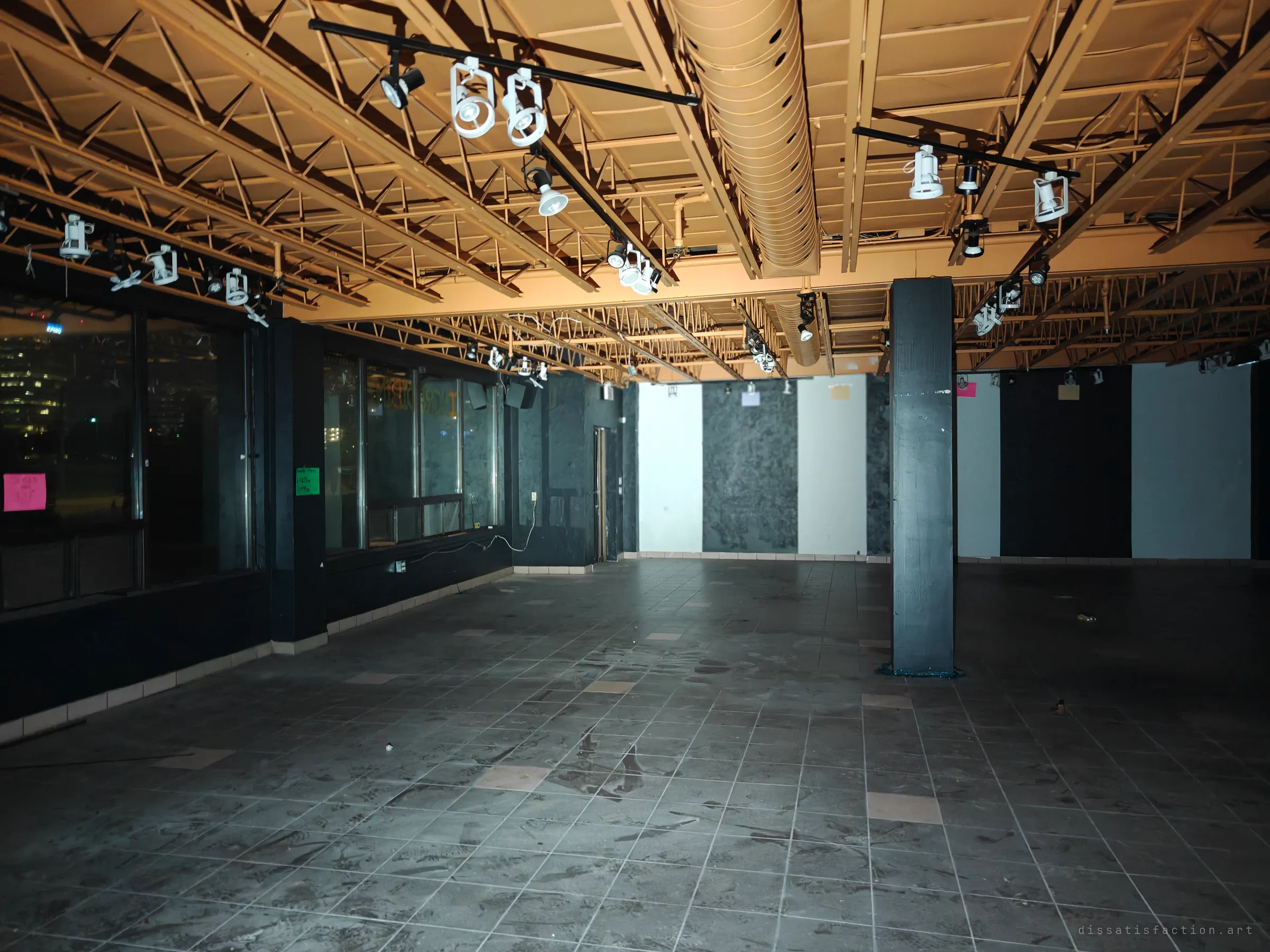 A large dark retail hall under an exposed orange-painted truss ceiling, still strung with track spotlights above a dusty tiled floor. Faded notices hang in the windows, with city lights showing beyond.