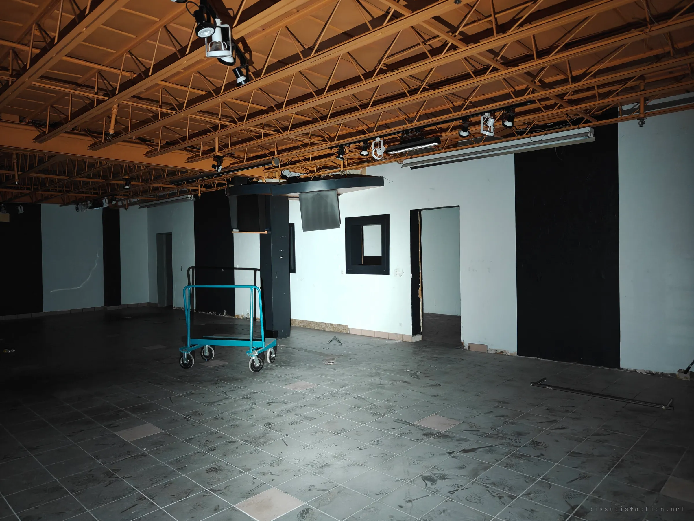 The same hall seen from another angle: black and white wall panels, a service window and an empty teal flatbed cart on a footprint-covered tile floor. Track lights still hang from the open trusses overhead.