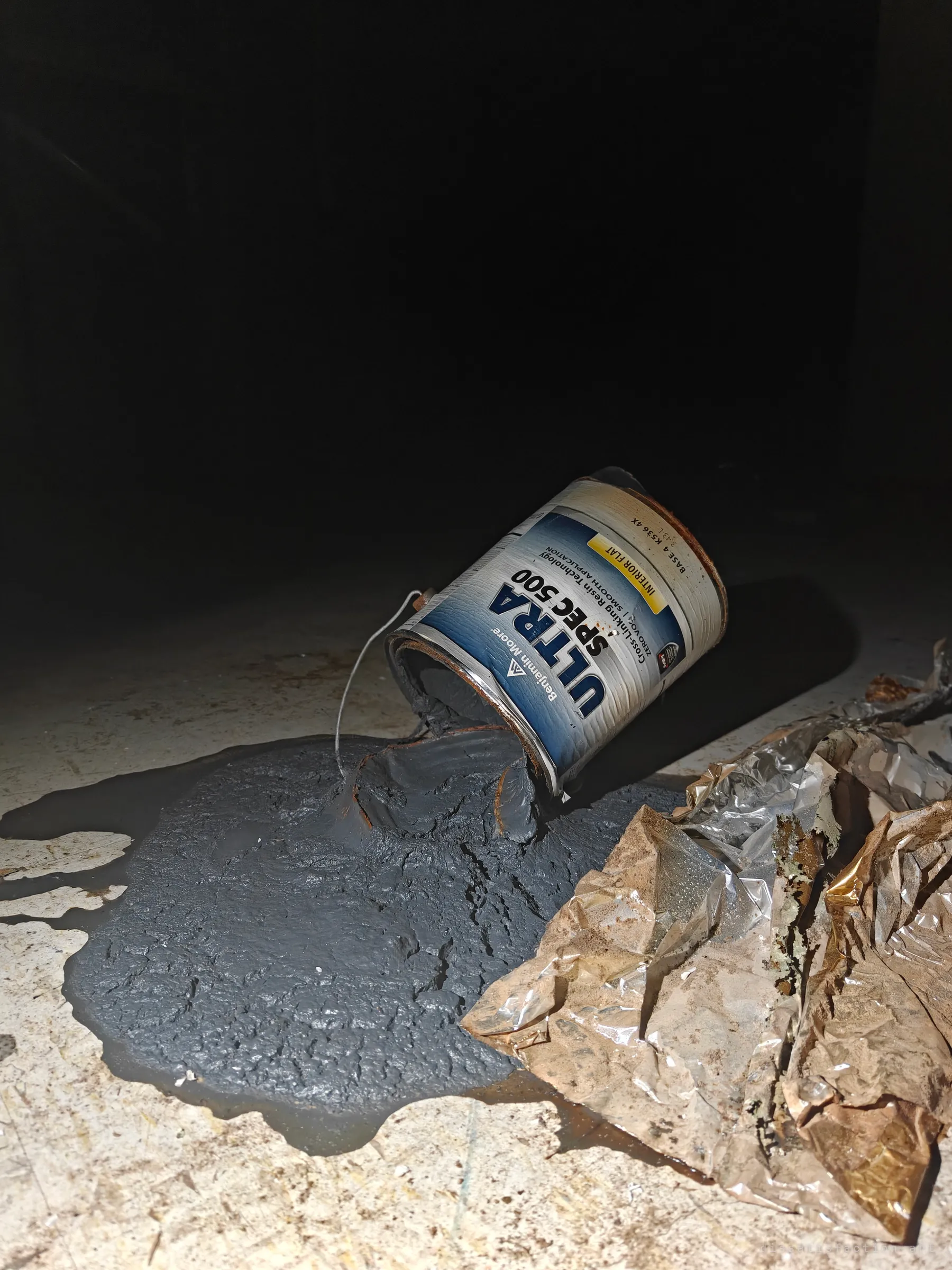 A tipped paint can has spilled a thick grey pool across a dusty floor beside crumpled foil. The spill is lit by flashlight against total darkness.