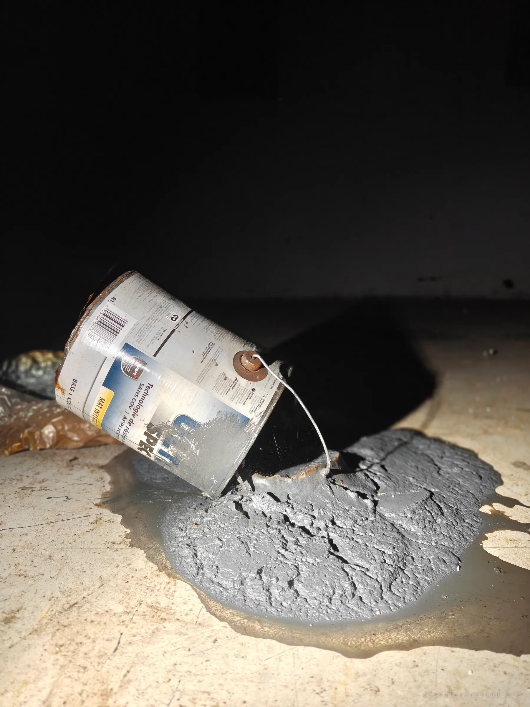 A rusted paint can lies tipped on its side, its grey contents spilled and set into a cracked puddle on the pale floor. A small still life of a job abandoned mid-way.