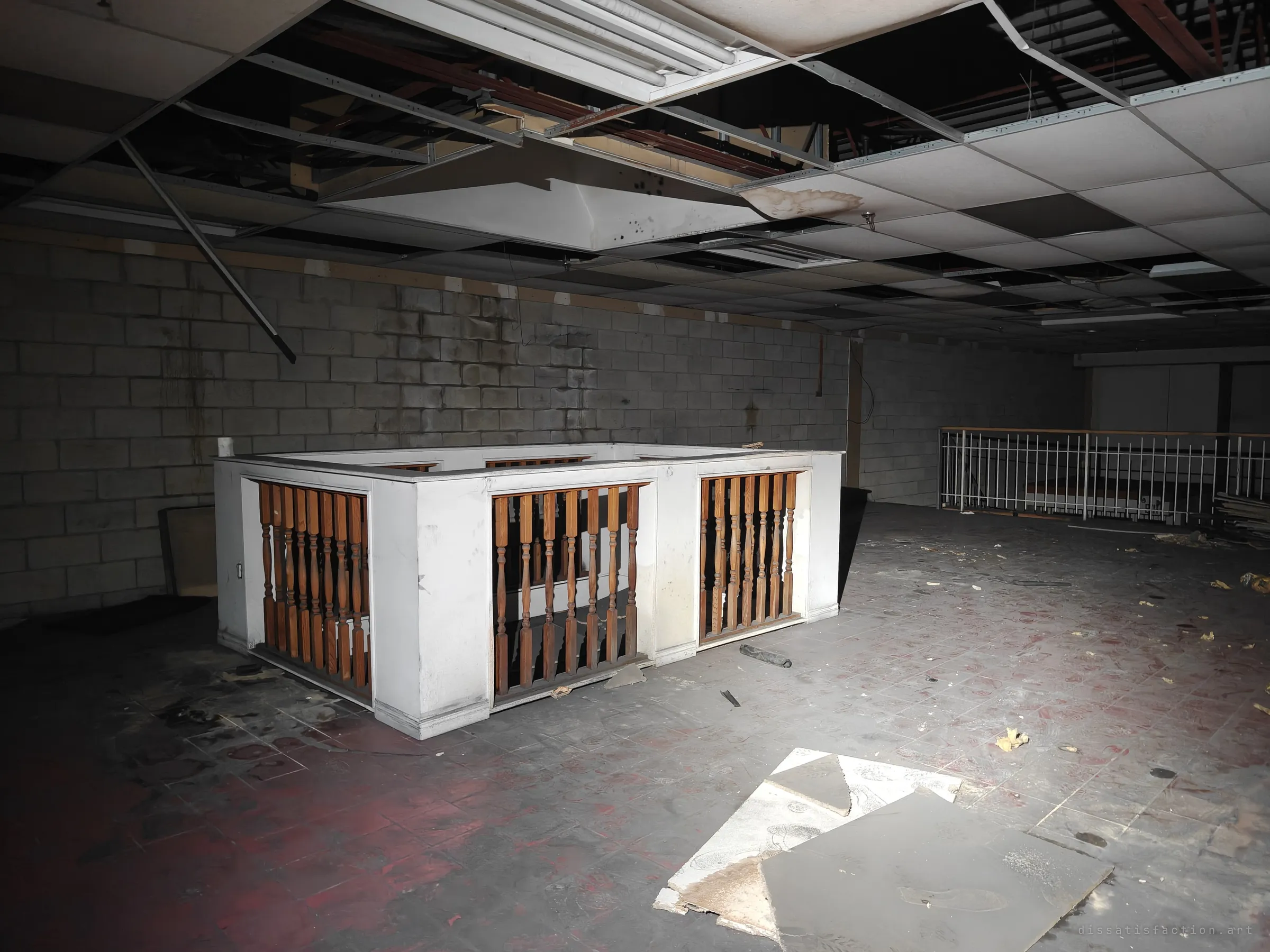 A white stairwell enclosure with turned wooden balusters stands in a dark upper floor under a half-collapsed drop ceiling. Loose ceiling tiles lie on the stained floor.