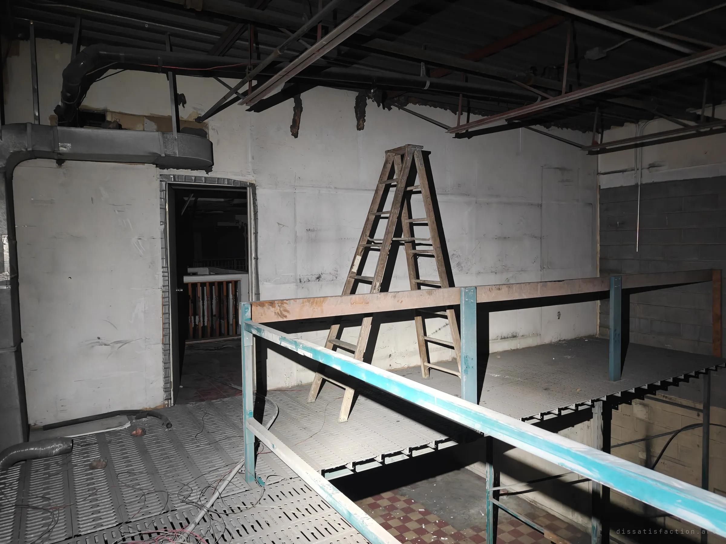 A wooden stepladder stands on a perforated steel mezzanine beneath exposed ducts and joists, a doorway beyond showing the wooden balustrade of a stairwell. Loose wiring snakes across the grating.