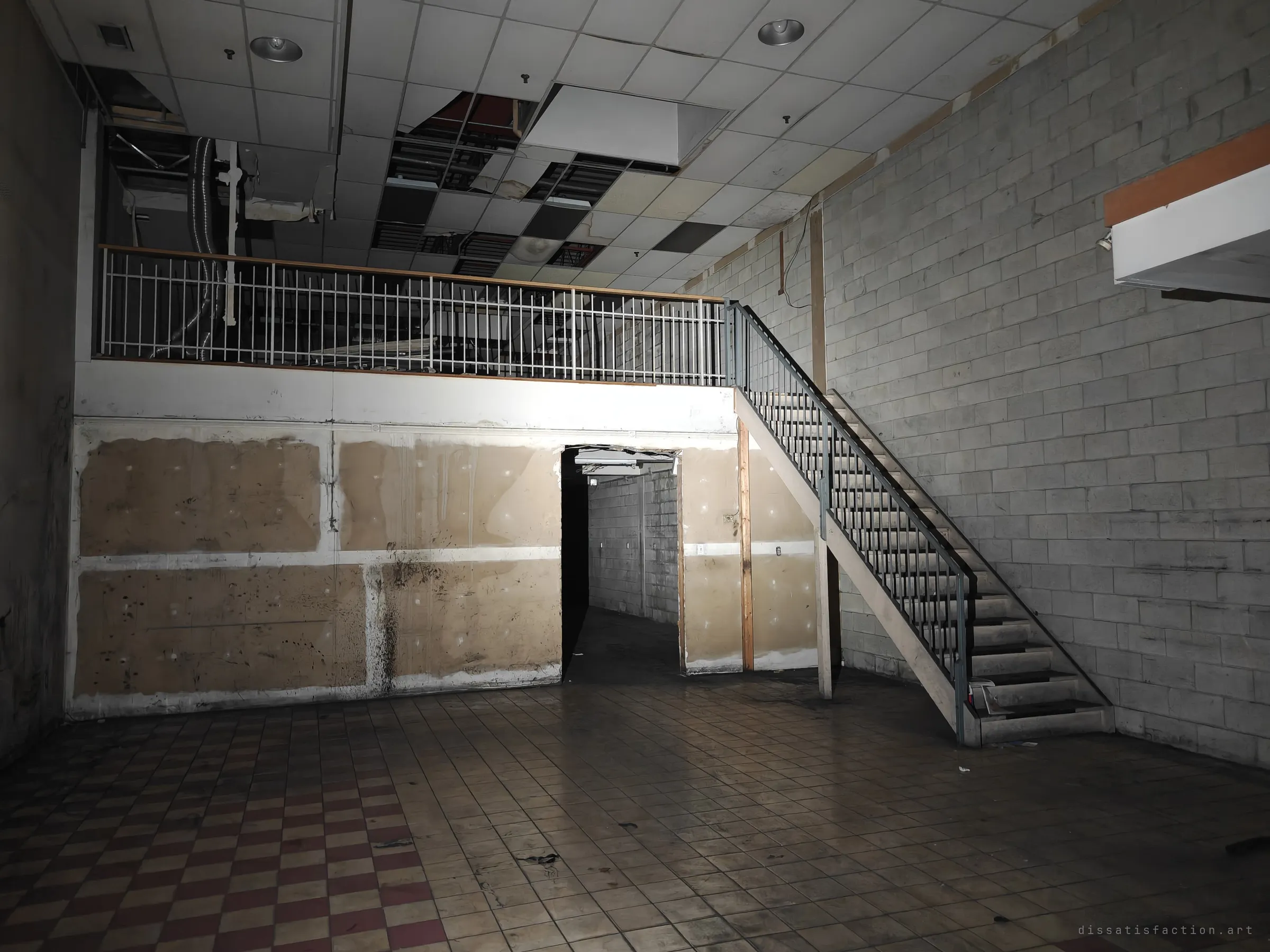 A stripped shop floor with a steel stair climbing to a railed mezzanine, a narrow corridor leading off beneath it. Missing ceiling tiles leave dark gaps overhead.