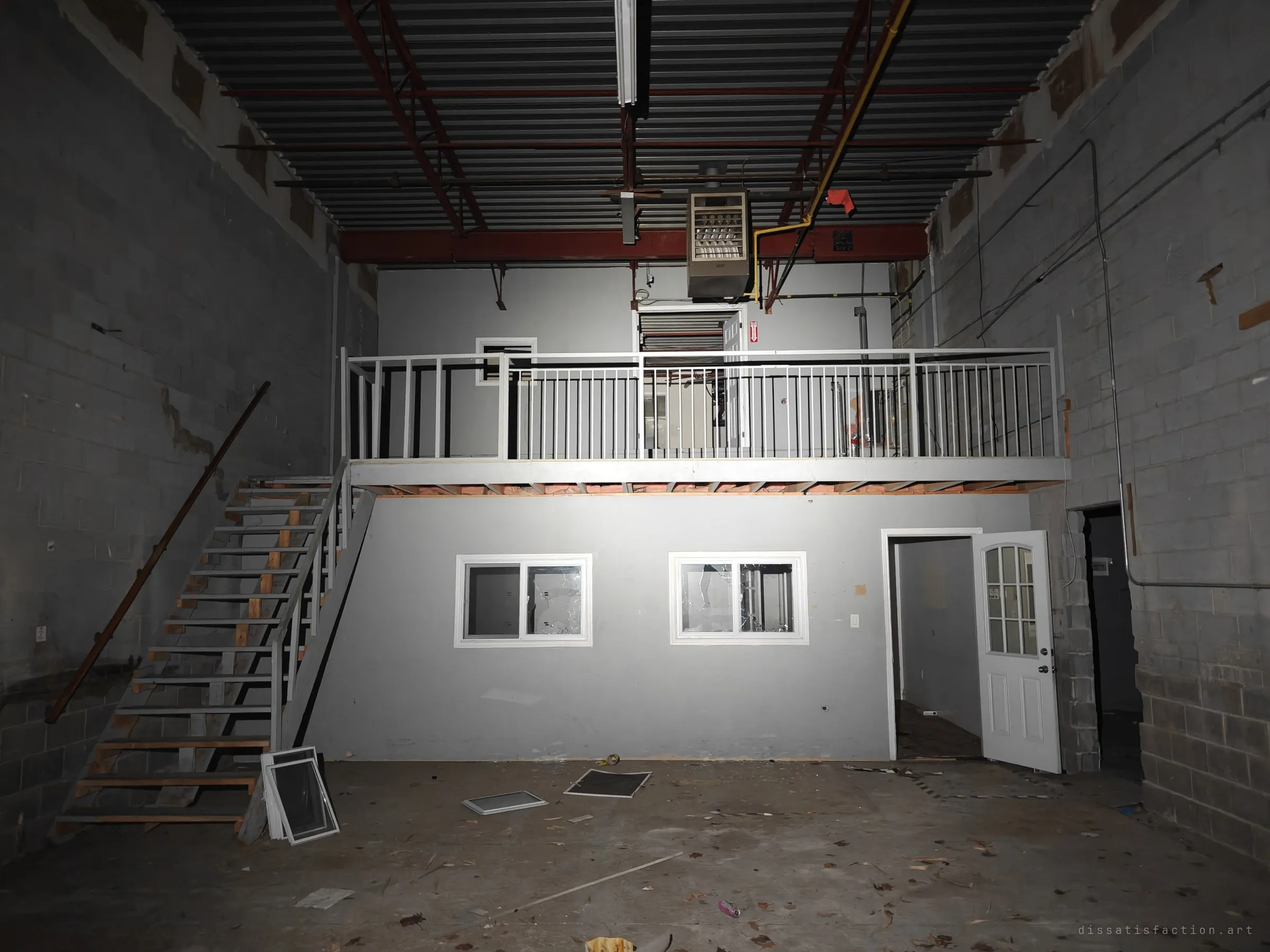A tall block-walled unit with a two-level office built inside it: smashed windows below, a railed balcony above and a stair on the left. Window frames lie scattered on the concrete.