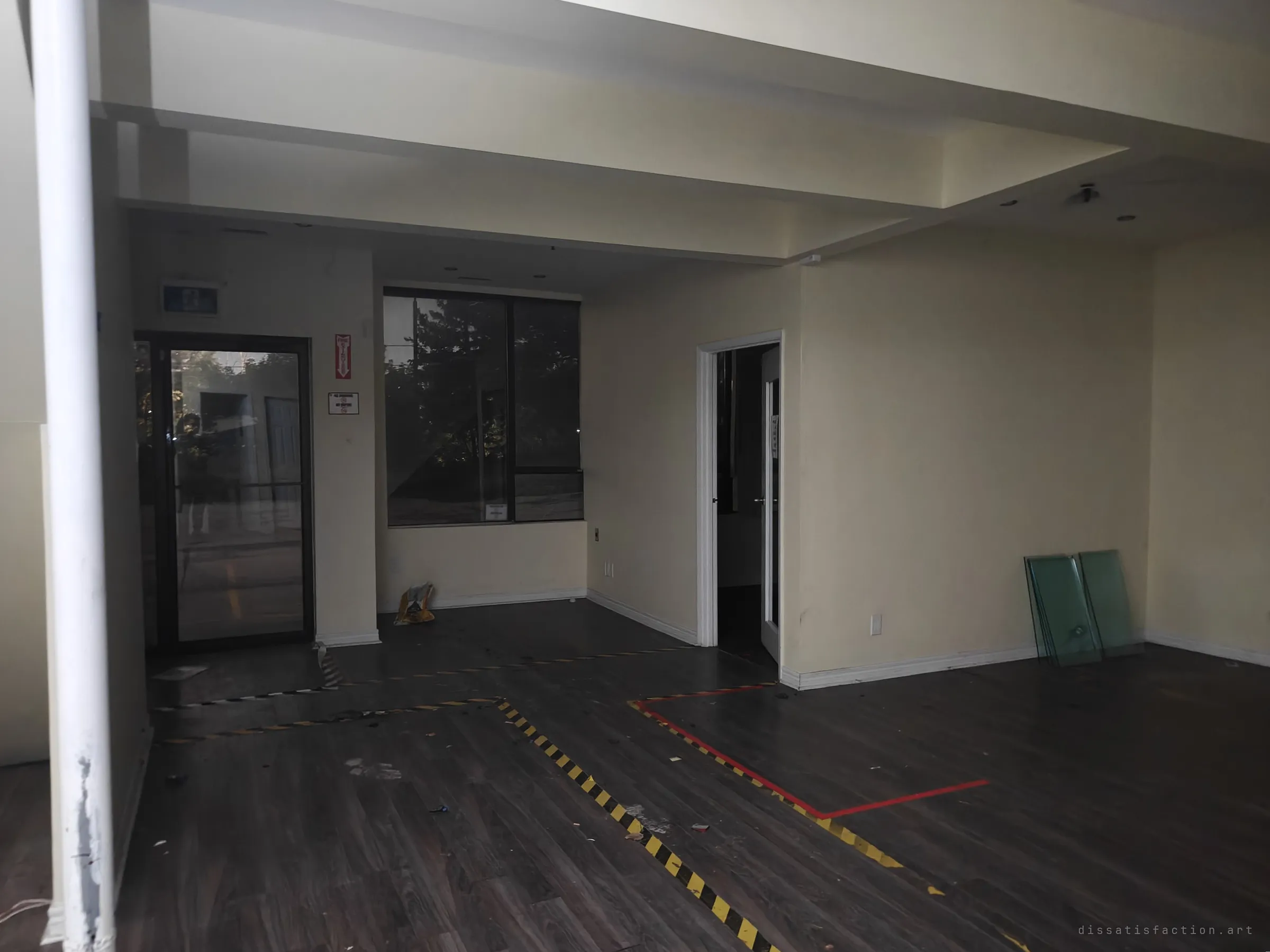 An emptied front unit with dark laminate floor still marked out in yellow-black and red tape, glass panes leaning on the wall. Evening light comes through the glass door and window, the last trace of a storefront.