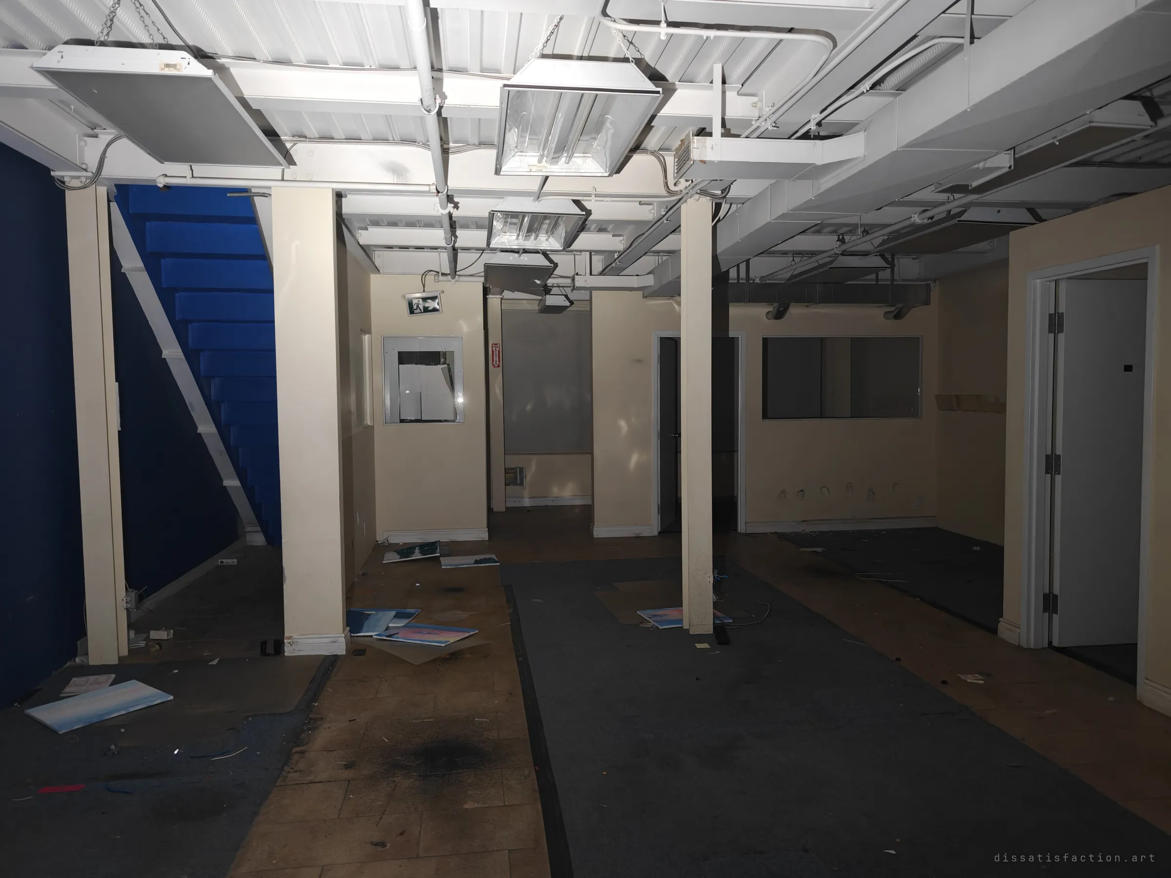 A gutted back office under exposed white-painted ceiling steel, a bright blue stairwell on the left. Abandoned canvases lie scattered across the stained floor.