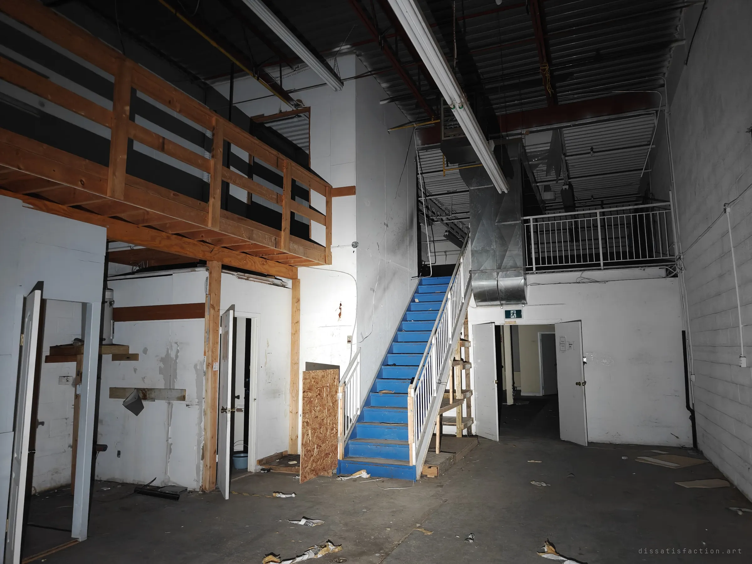 A tall industrial unit with a timber mezzanine on one side and a blue-painted staircase climbing to a steel-railed loft. Open doors lead back into the office rooms.