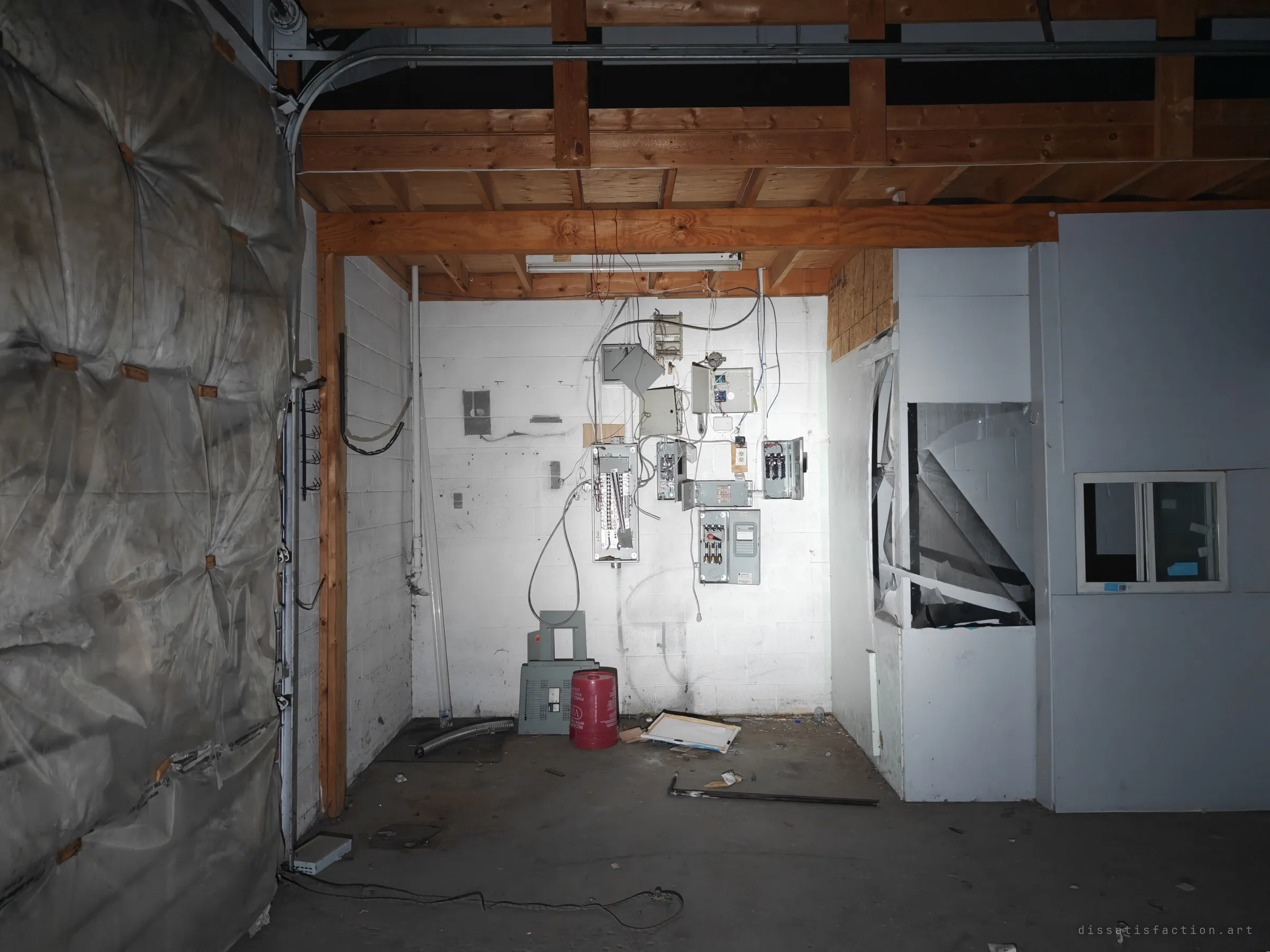 A corner of the warehouse where stripped electrical panels hang from a white block wall under timber joists. A plastic-wrapped roll-up door fills the left side of the frame.