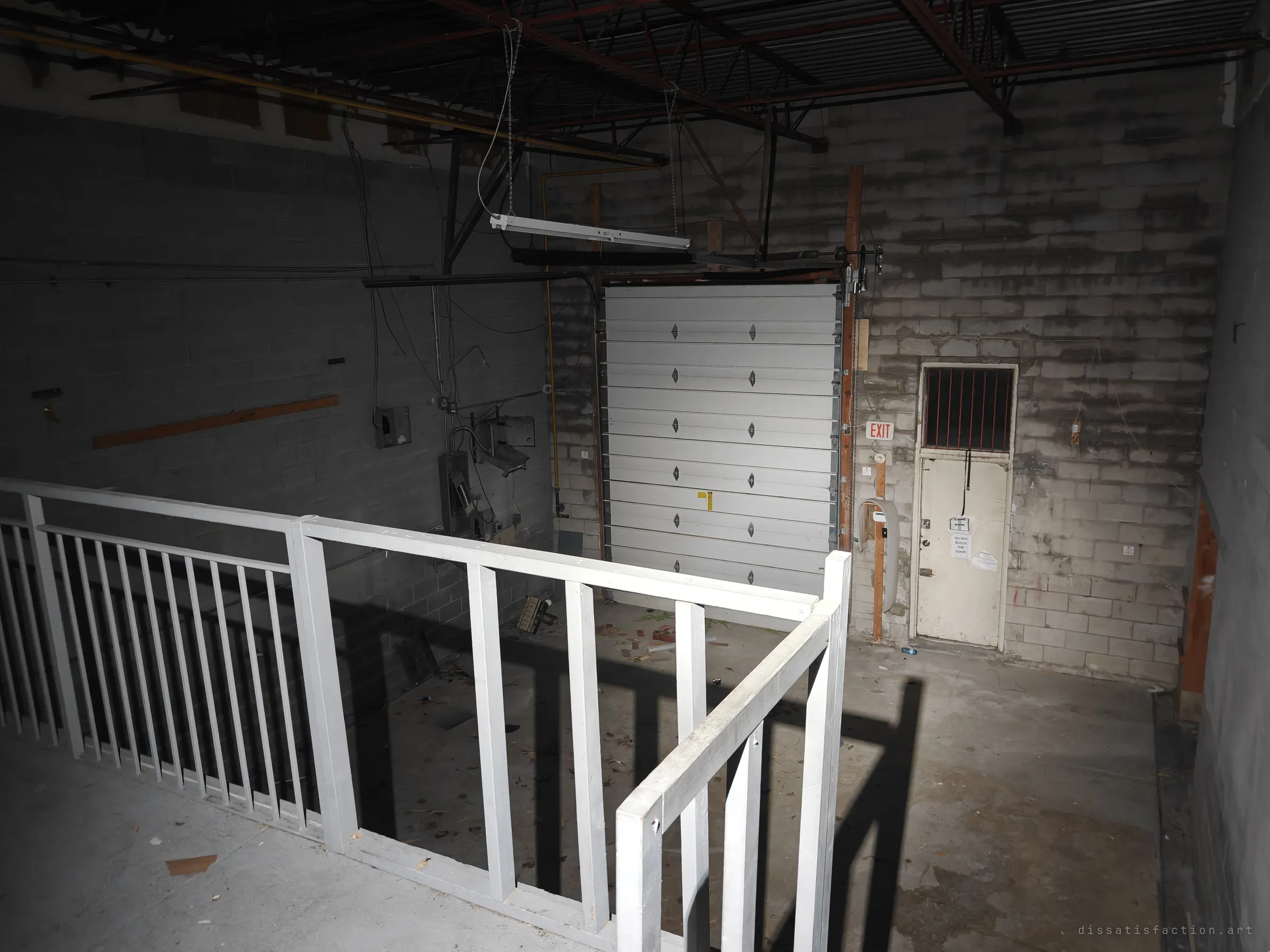 Looking down from the mezzanine railing onto a bare loading bay with a white roll-up door and a barred service door. A flashlight throws the railing shadows across the concrete.