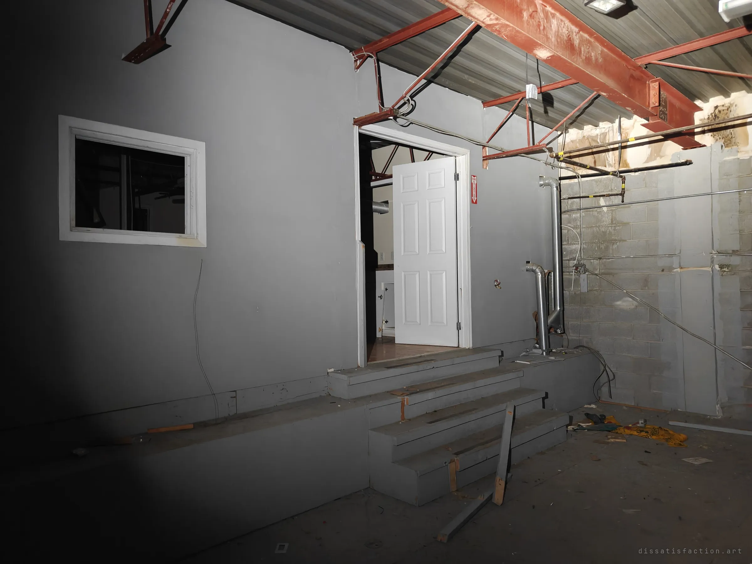The outside of a grey-painted office box built inside the warehouse, with steps up to an open door. Red steel beams and a rusted roof deck hang overhead.