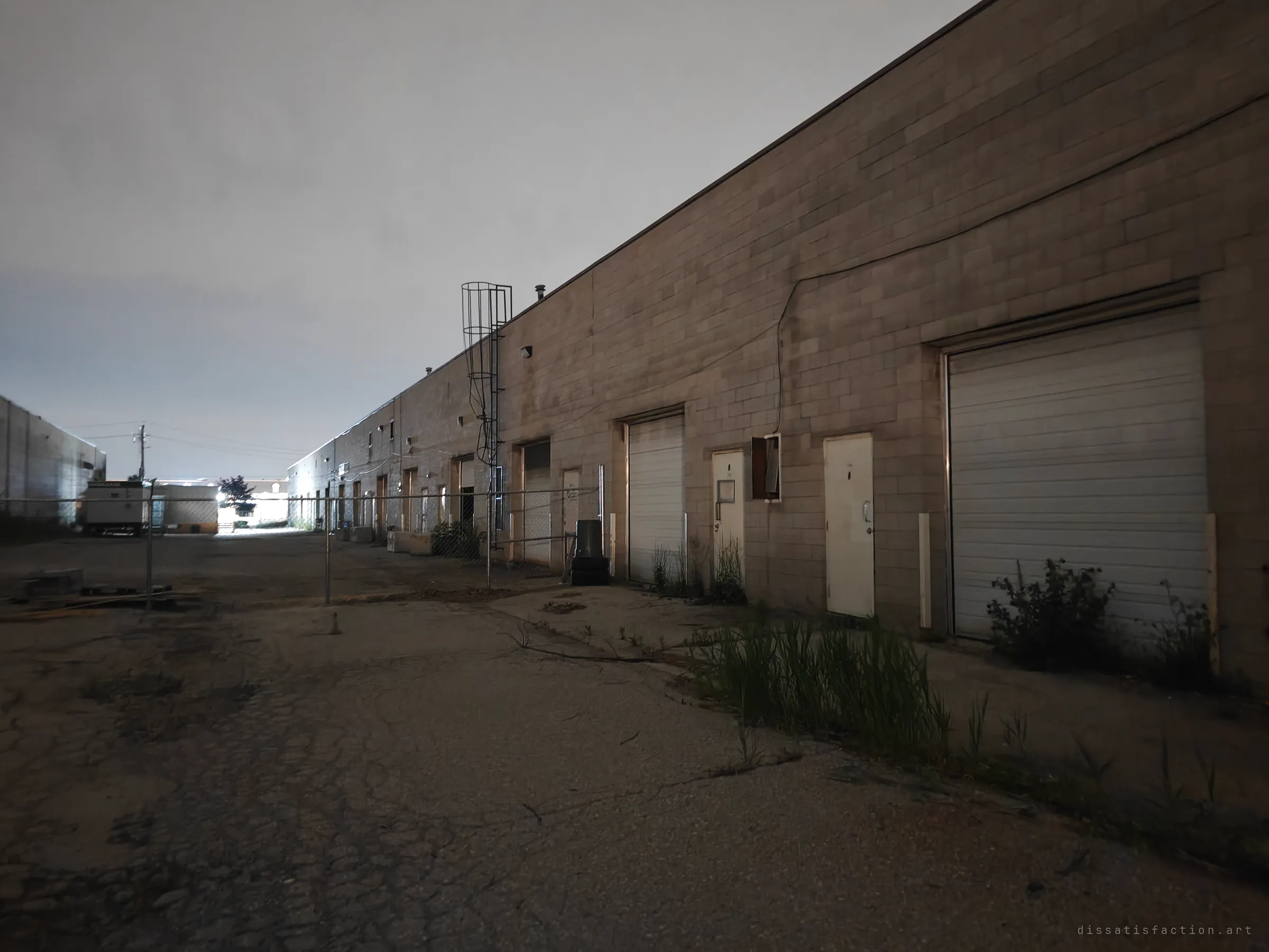 The back of a long block industrial row at night, its roll-up doors and steel doors lined up under a hazy sky. Weeds push through the cracked asphalt of the empty lot.