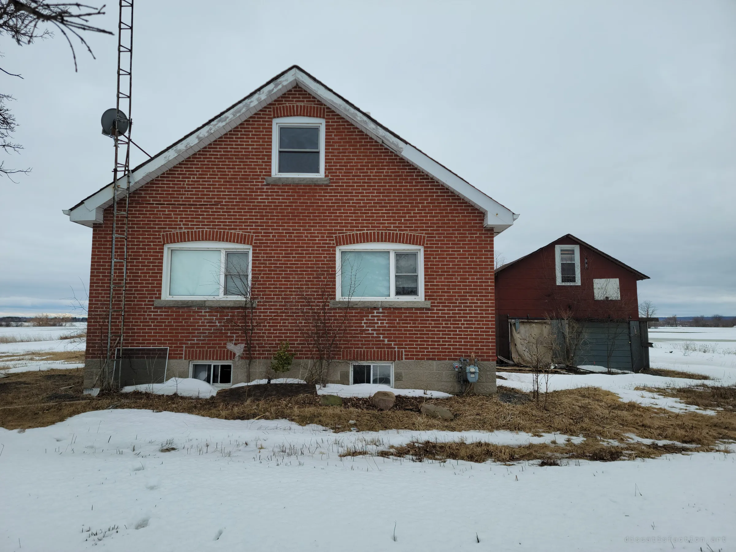 The red-brick gable end of an abandoned farmhouse sits in thawing snow, a tall antenna mast at its side and a weathered red outbuilding next door. It sets the scene: a rural house left standing almost intact.