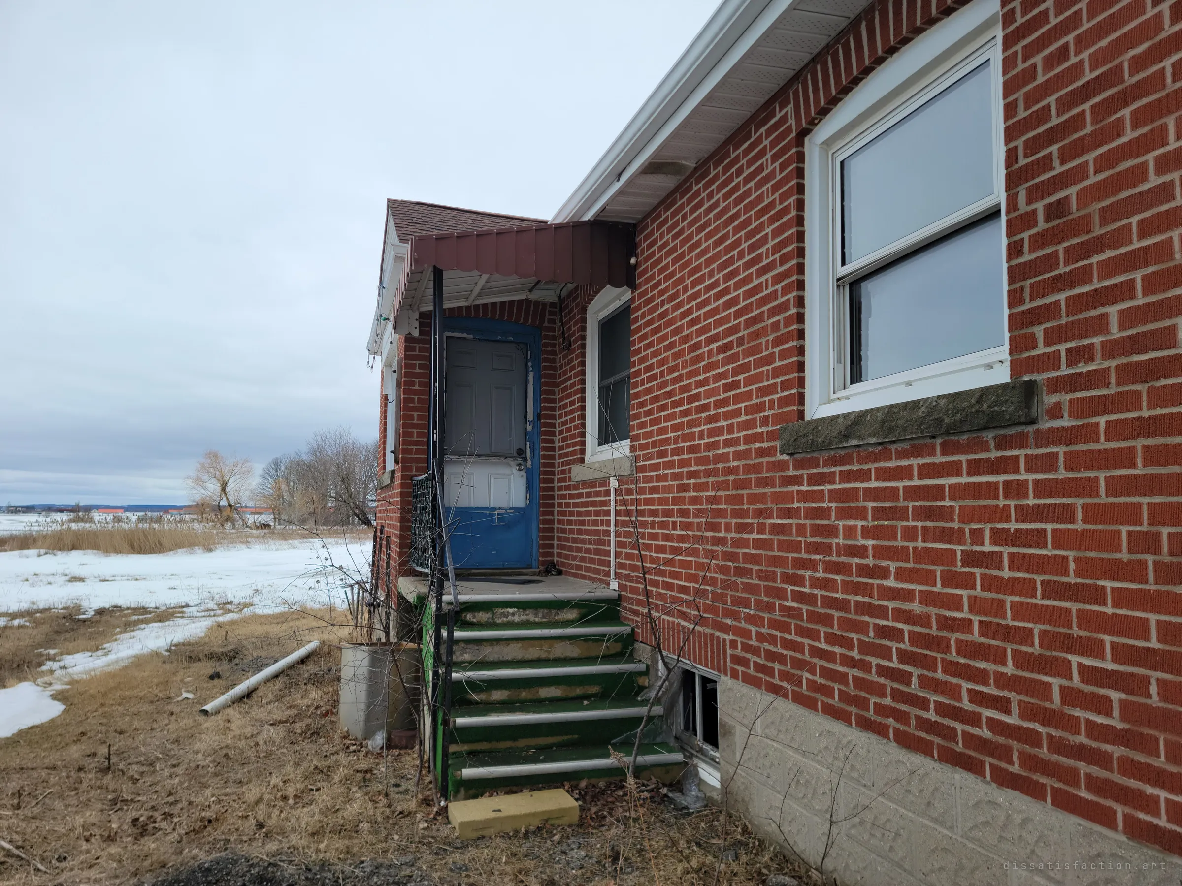 A side entrance with a blue storm door under a small metal awning, reached by green-carpeted steps along the brick wall. Snowy marsh and bare trees stretch away behind it.