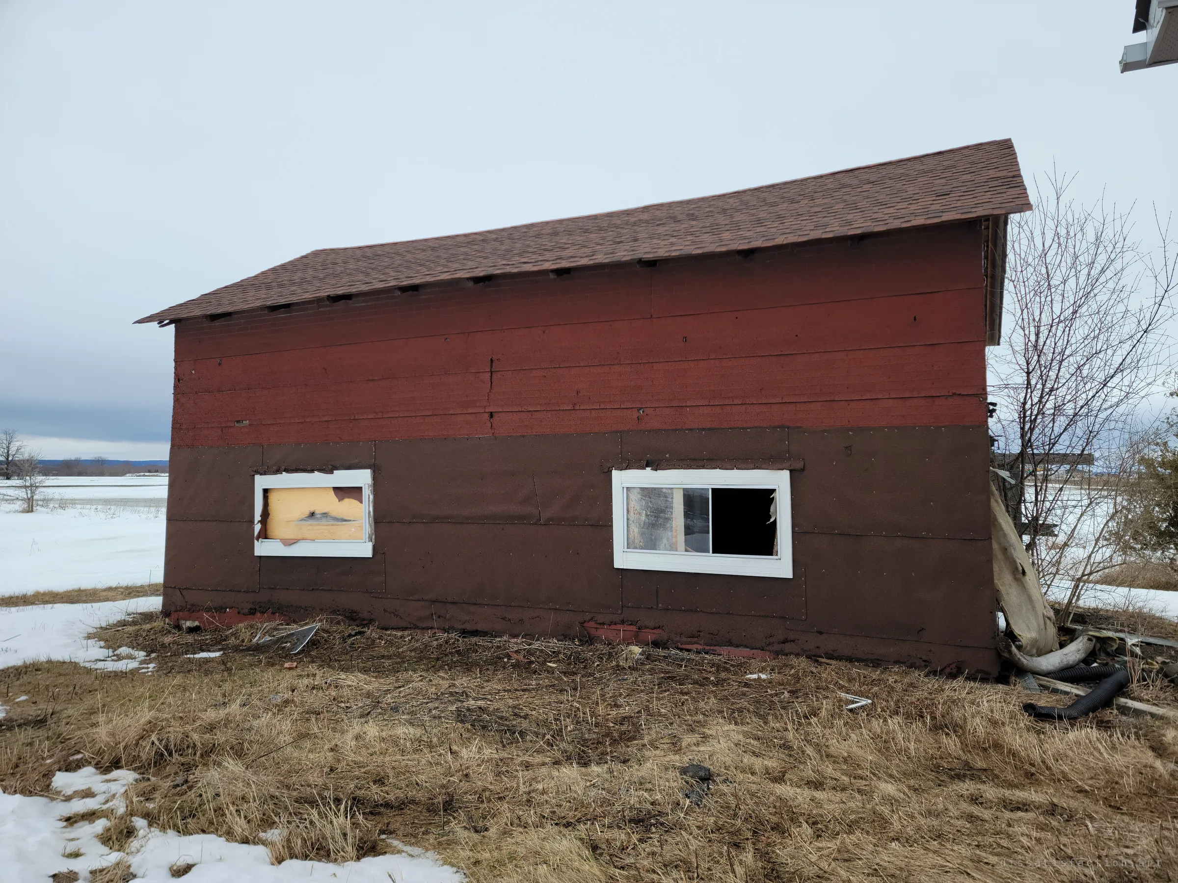 A sagging outbuilding clad in red and brown tar paper, one window boarded and the other broken, stands on flattened winter grass. Its bowed roofline shows years of neglect.