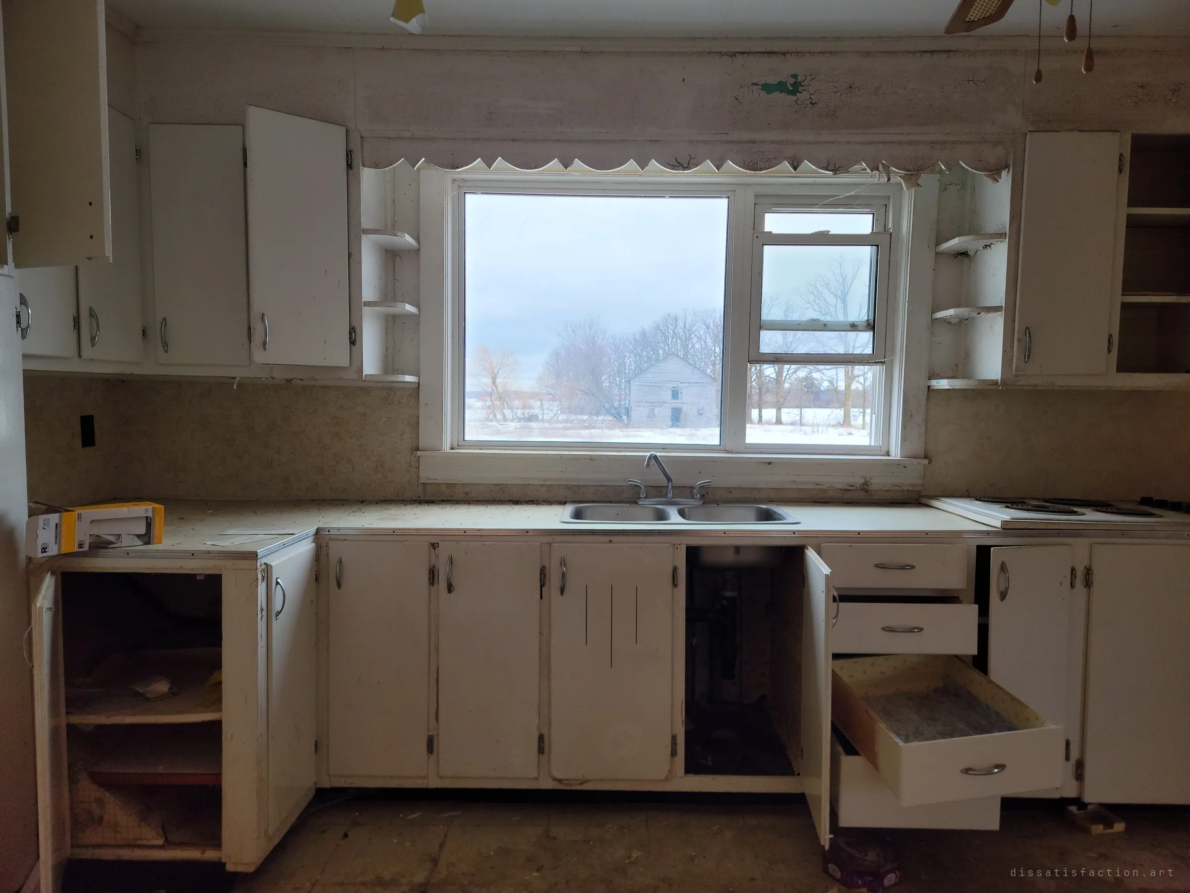 A white kitchen with doors and drawers hanging open frames a large window looking out on a grey barn in the snow. The scalloped valance and double sink are left just as they were.